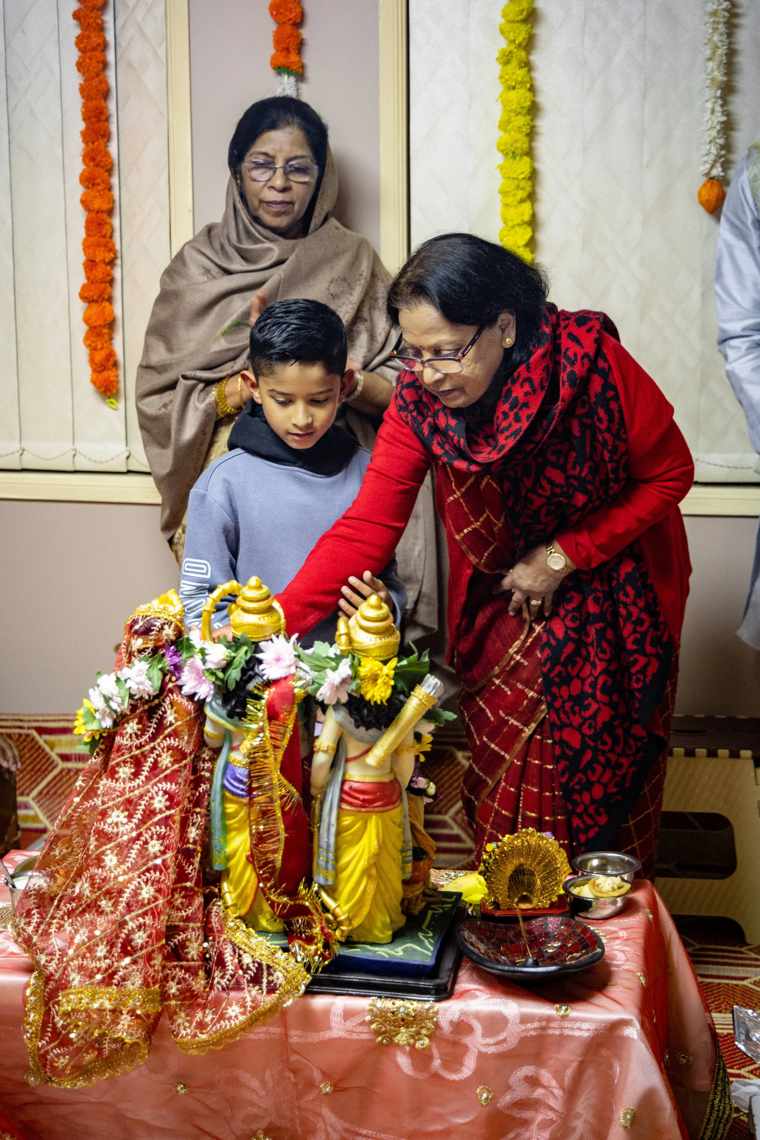 Two women and a young boy participating in a religious ceremony, touching a shrine with statues decorated with flowers and gold, surrounded by traditional items on a table.