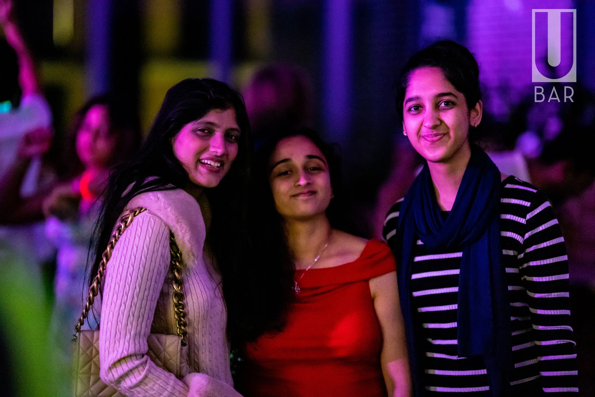 Three women smiling and posing together at a bar, with a purple and pink lit background.