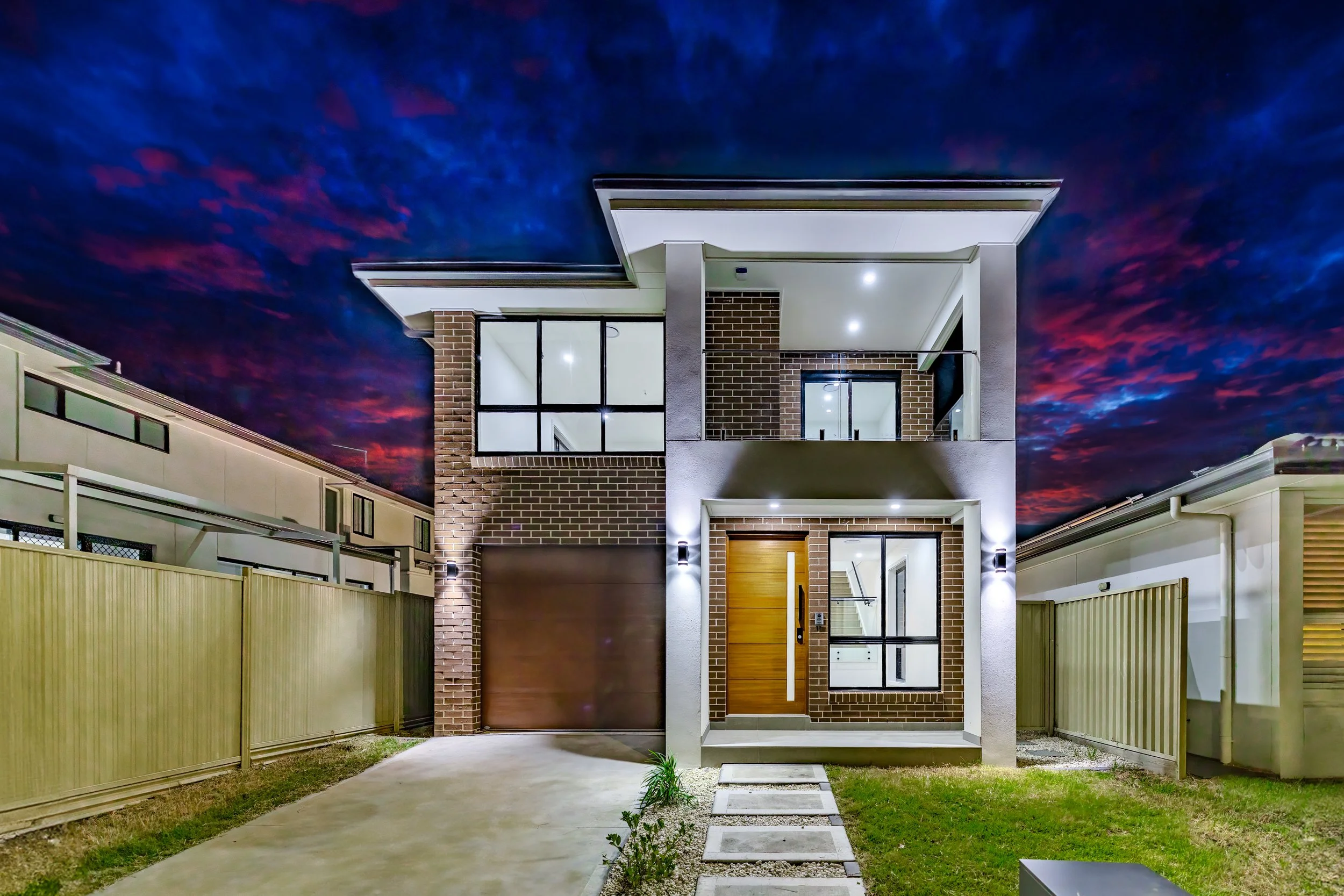 Modern two-story house illuminated at night, featuring brick and white exterior with large windows and a wooden front door, surrounded by a small lawn and paved pathway.