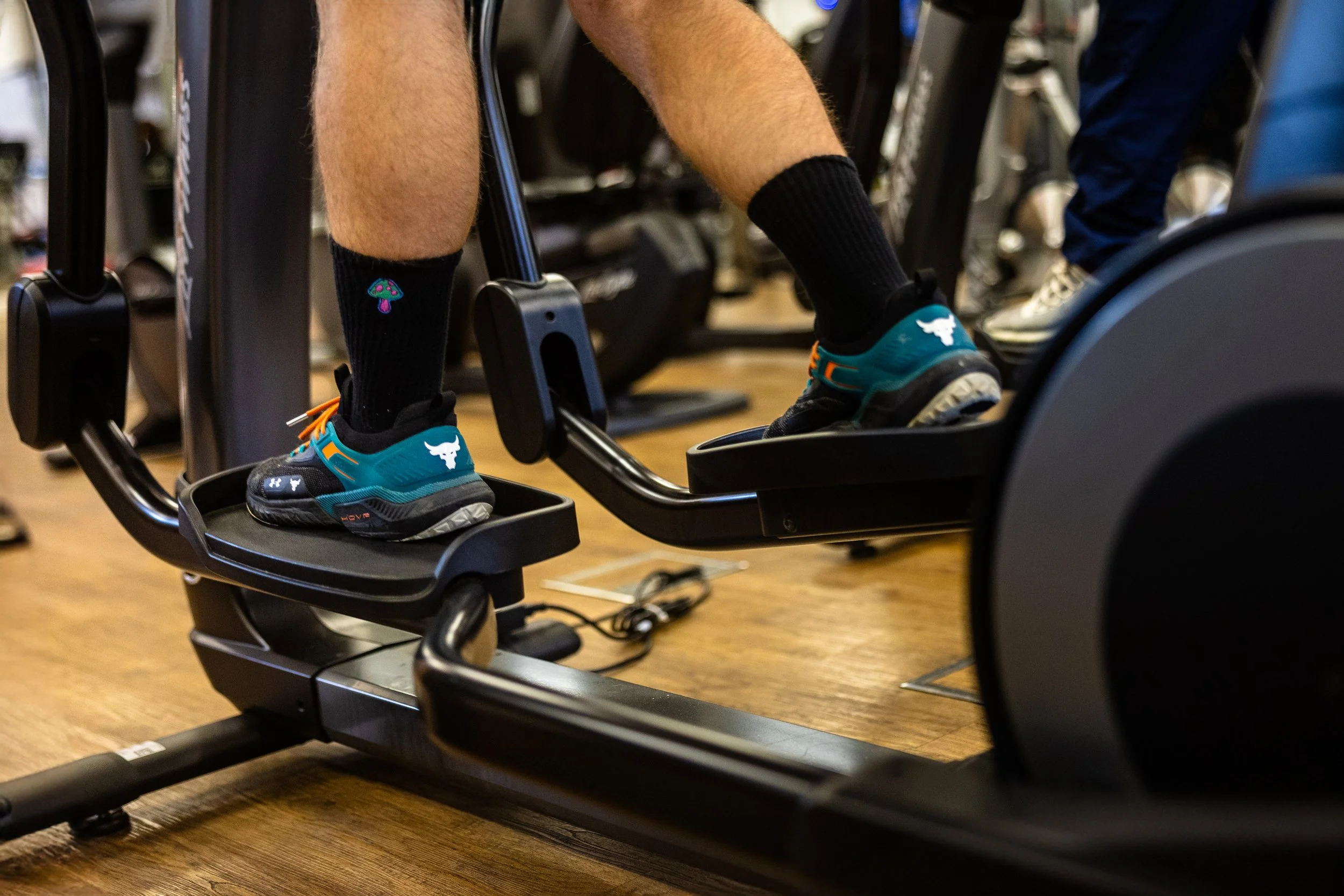 Close-up of a person using a rowing machine in a gym, wearing black socks with colorful mushrooms and teal athletic shoes with a bull logo.