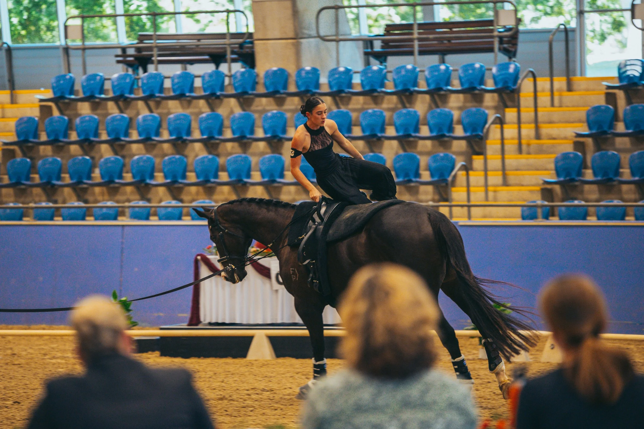 A woman dressed in black performing a dressage routine on a dark horse inside an indoor arena with blue seats and spectators watching.