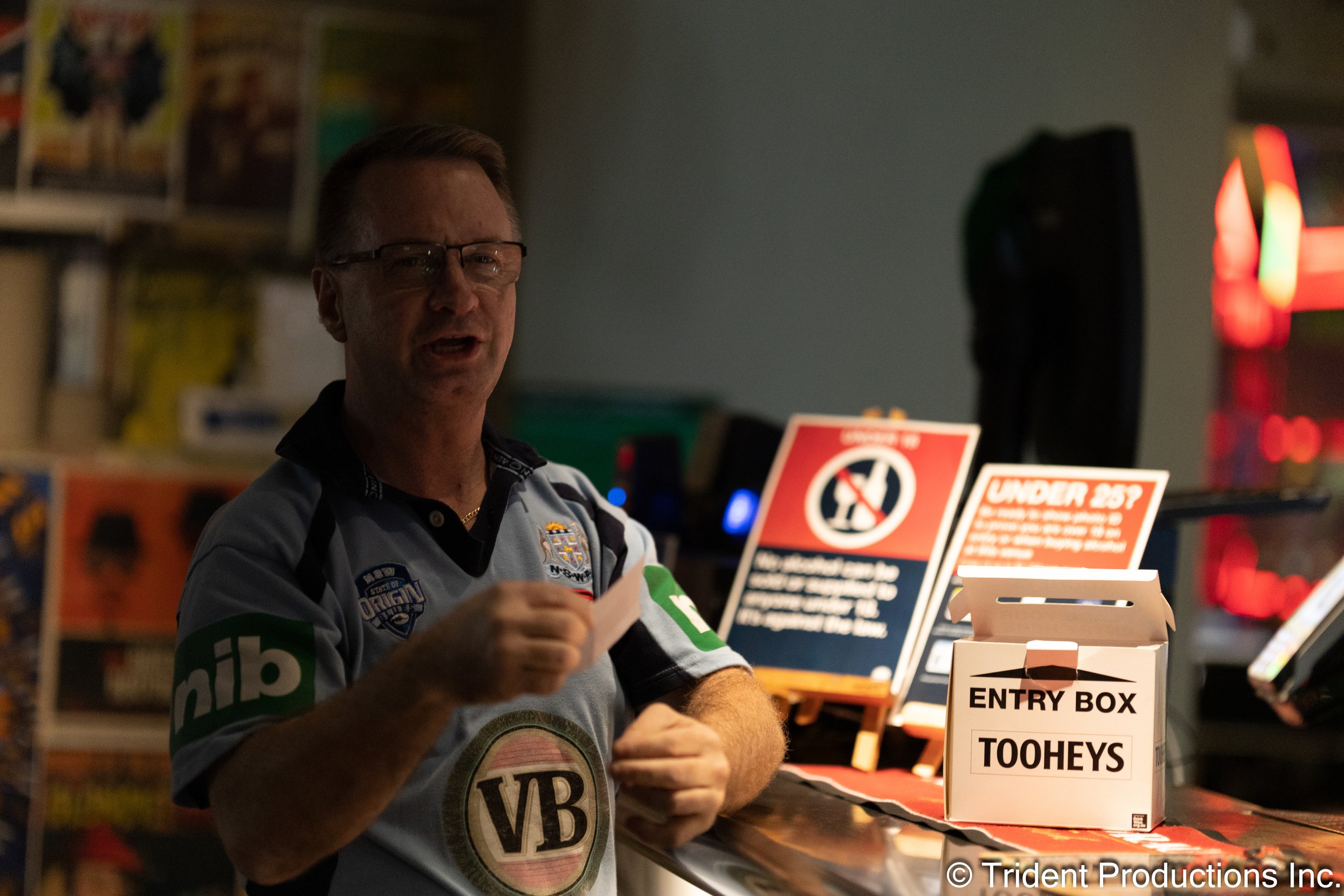 A man wearing glasses and a gray sports jersey sitting at a table, with a sign behind him that says 'Under 25?' and an entry box labeled 'TOOHEYS'.
