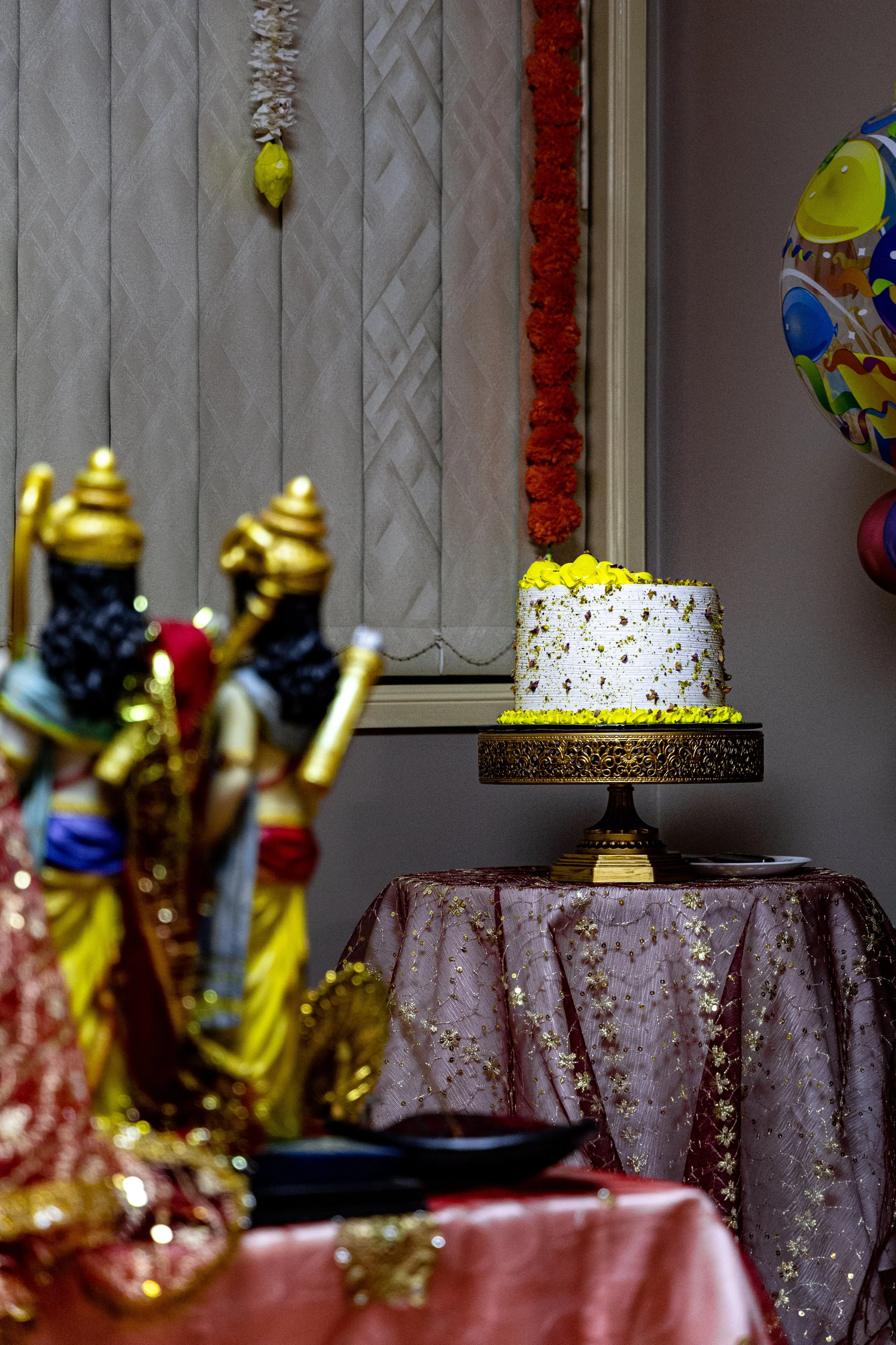 A white cake with yellow decoration on top, placed on a decorative cake stand, set on a table with a pink tablecloth, at a celebration with balloons and festive decorations.