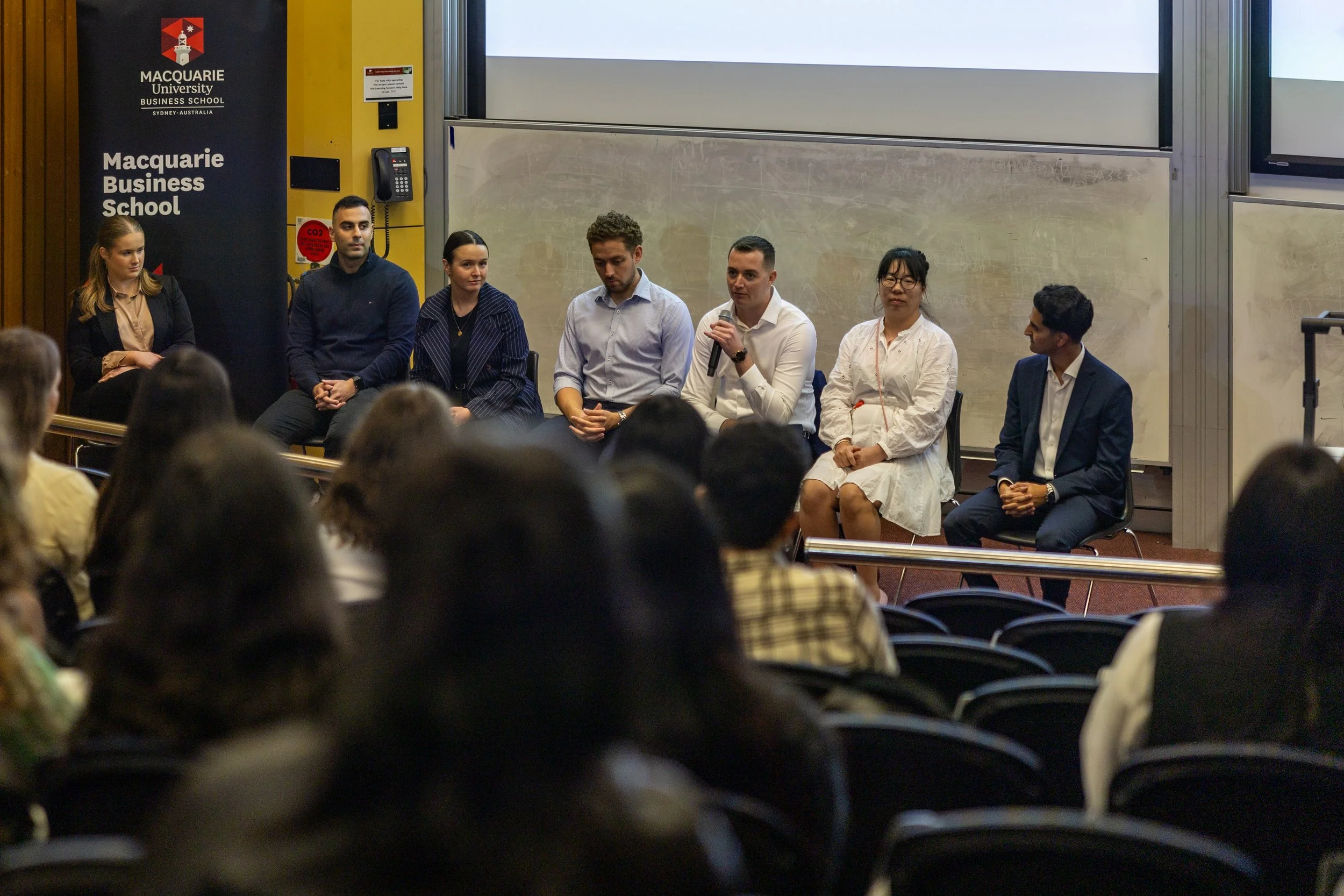 A panel of six diverse individuals sitting on chairs at the front of a conference room, with an audience in front of them. A Macquarie University Business School banner is visible on the left. One person is speaking into a microphone.