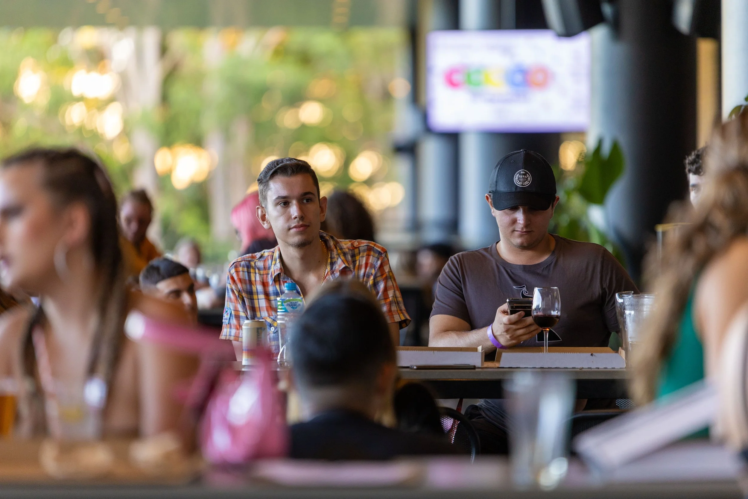 People sitting at tables in a lively indoor restaurant or bar, with one young man in a checkered shirt looking to the side and another in a dark shirt with a cap looking at their phone.