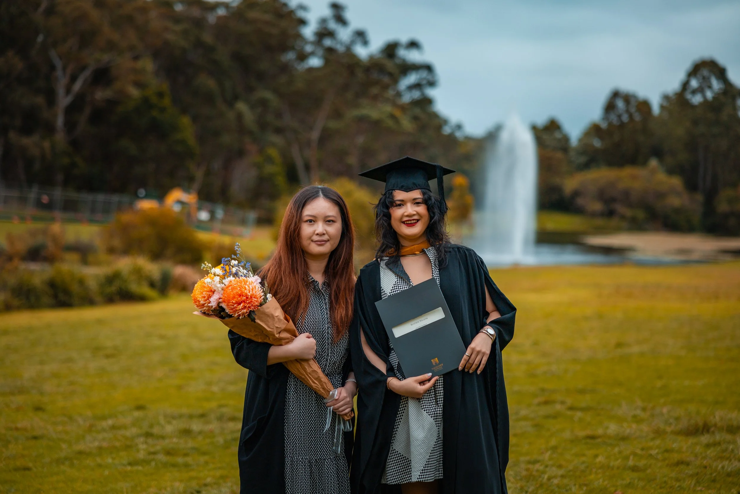 Two women in graduation gowns and caps standing outdoors on a grassy field with a fountain and trees in the background. One woman is holding a bouquet of flowers, and the other is holding a diploma.