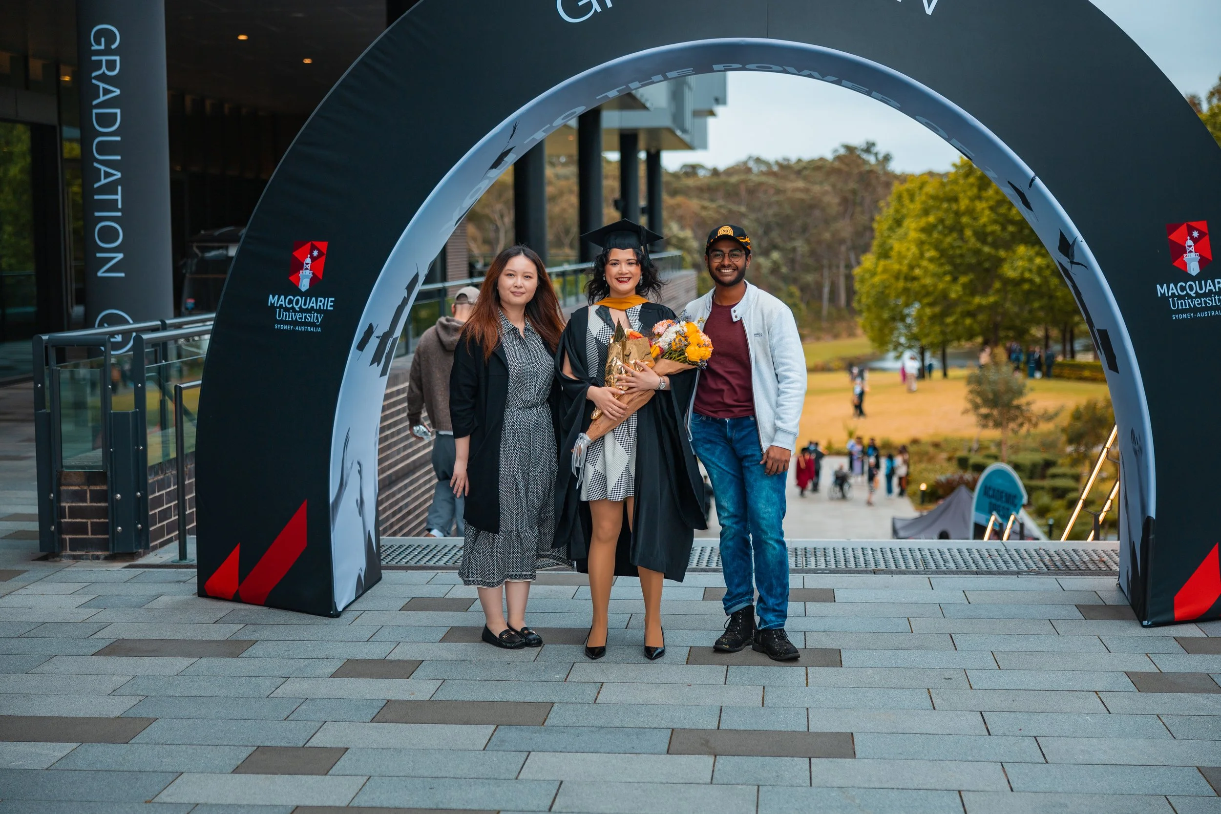 Graduation celebration at Macquarie University with three people standing under a large arch, one in graduation gown holding a bouquet, on outdoor campus with trees and other students in the background.