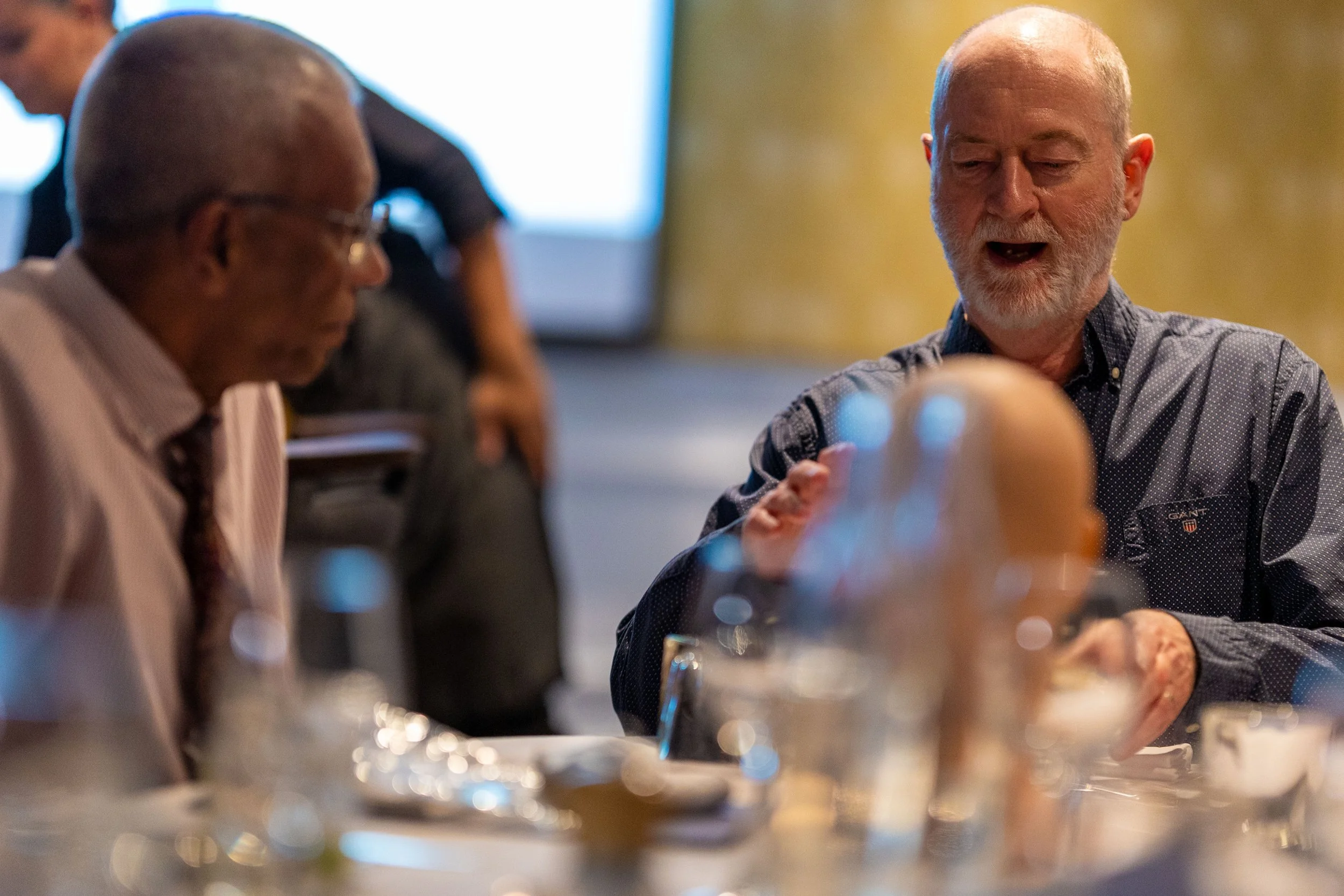 Two men sitting at a table in a social setting, engaging in a conversation, with blurred background and table items including glasses and bottles.