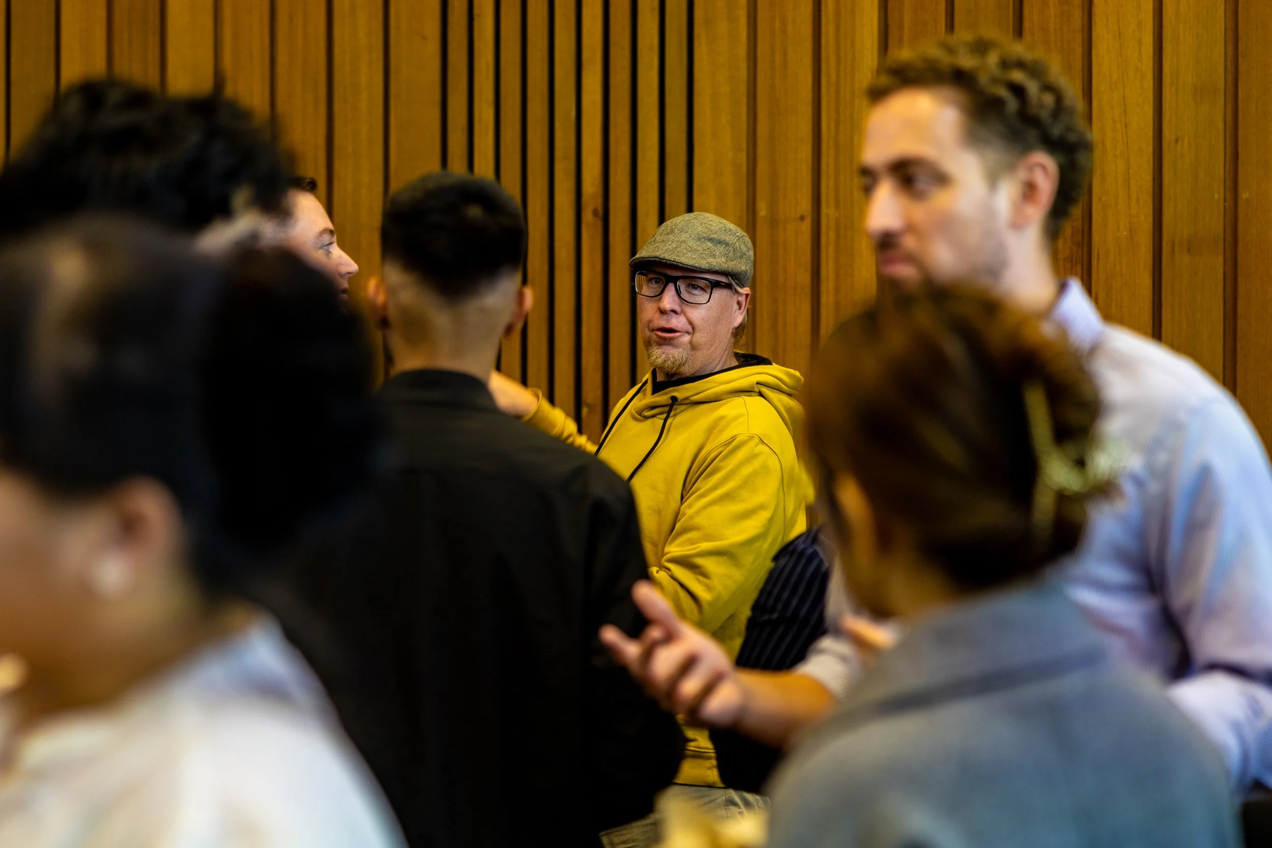 Man in a yellow hoodie and gray flat cap speaking to a group of diverse people in a room with wooden paneling.