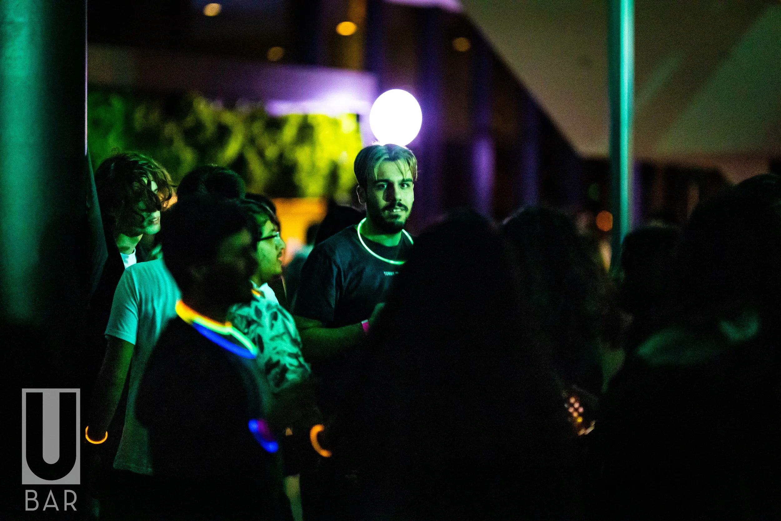 People at a nightclub wearing glow-in-the-dark accessories, illuminated by colorful lights, with one person looking at the camera.