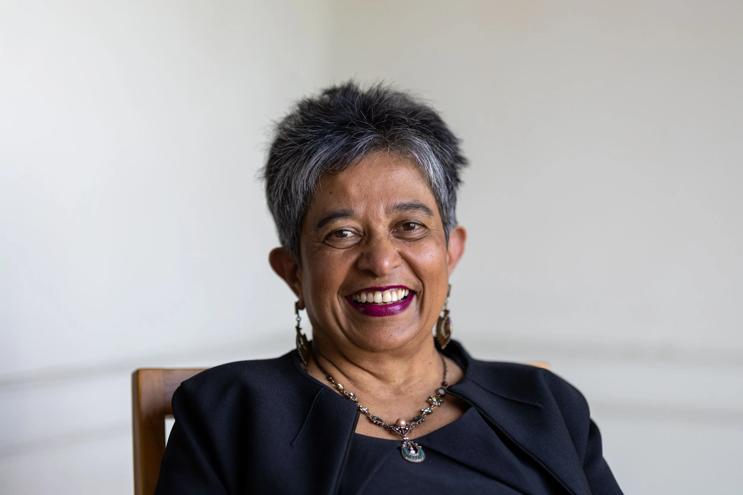 A smiling middle-aged woman with short, dark and gray hair, wearing a black top and a jewelry set with earrings, necklace, and pendant, sitting in front of a plain, light-colored wall.