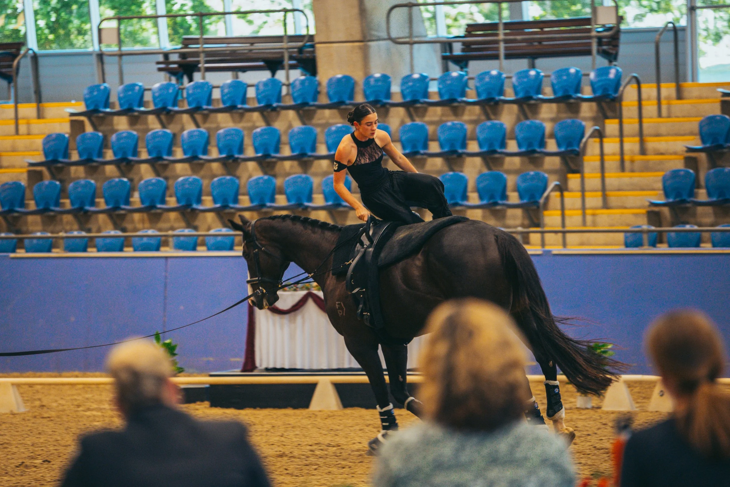 A woman in black clothing performing a dressage routine on a dark horse inside an indoor arena with blue seating and spectators watching.