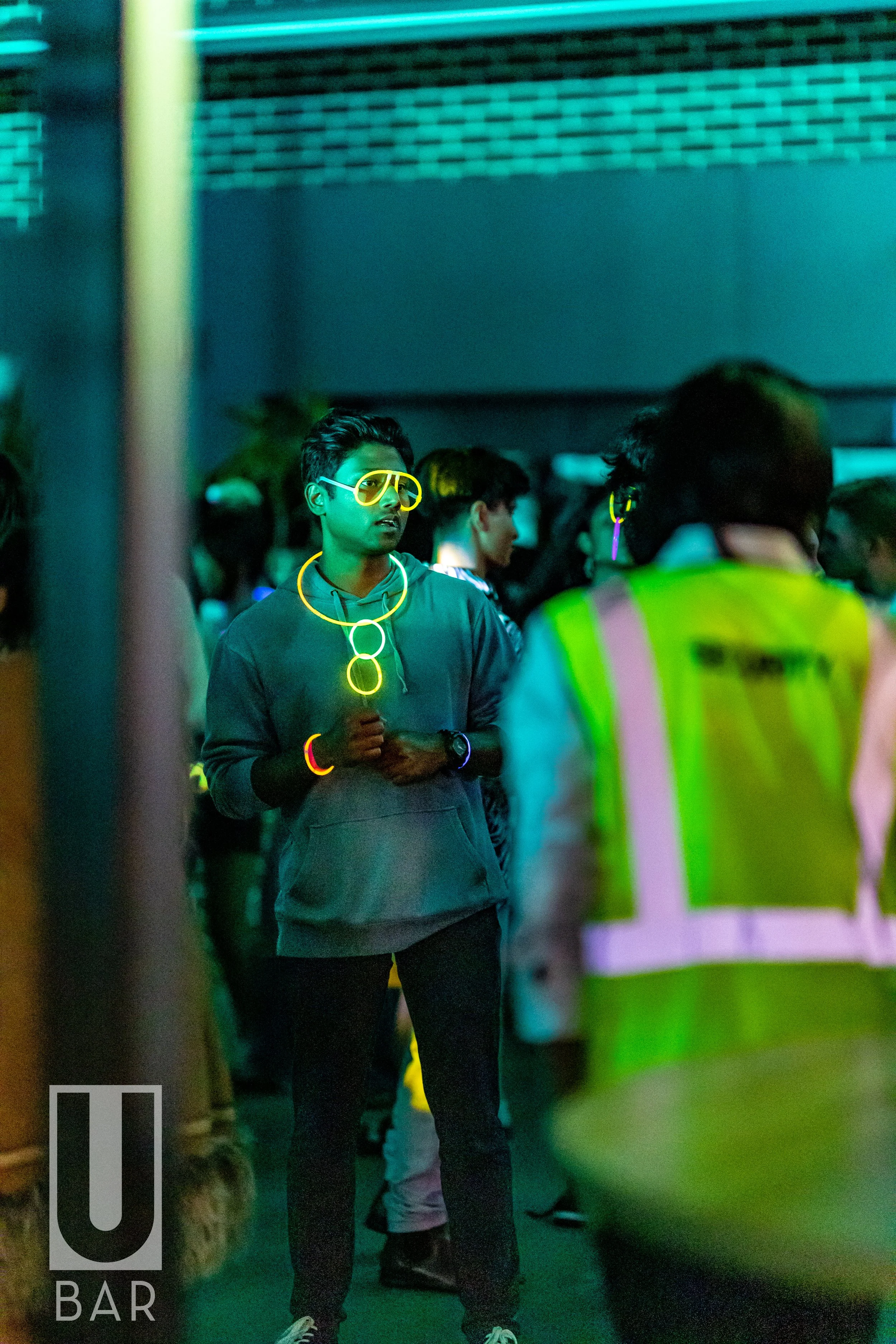 A young man at a neon-lit party wearing glow-in-the-dark jewelry and glasses, standing among a crowd with someone in a high-visibility vest nearby.