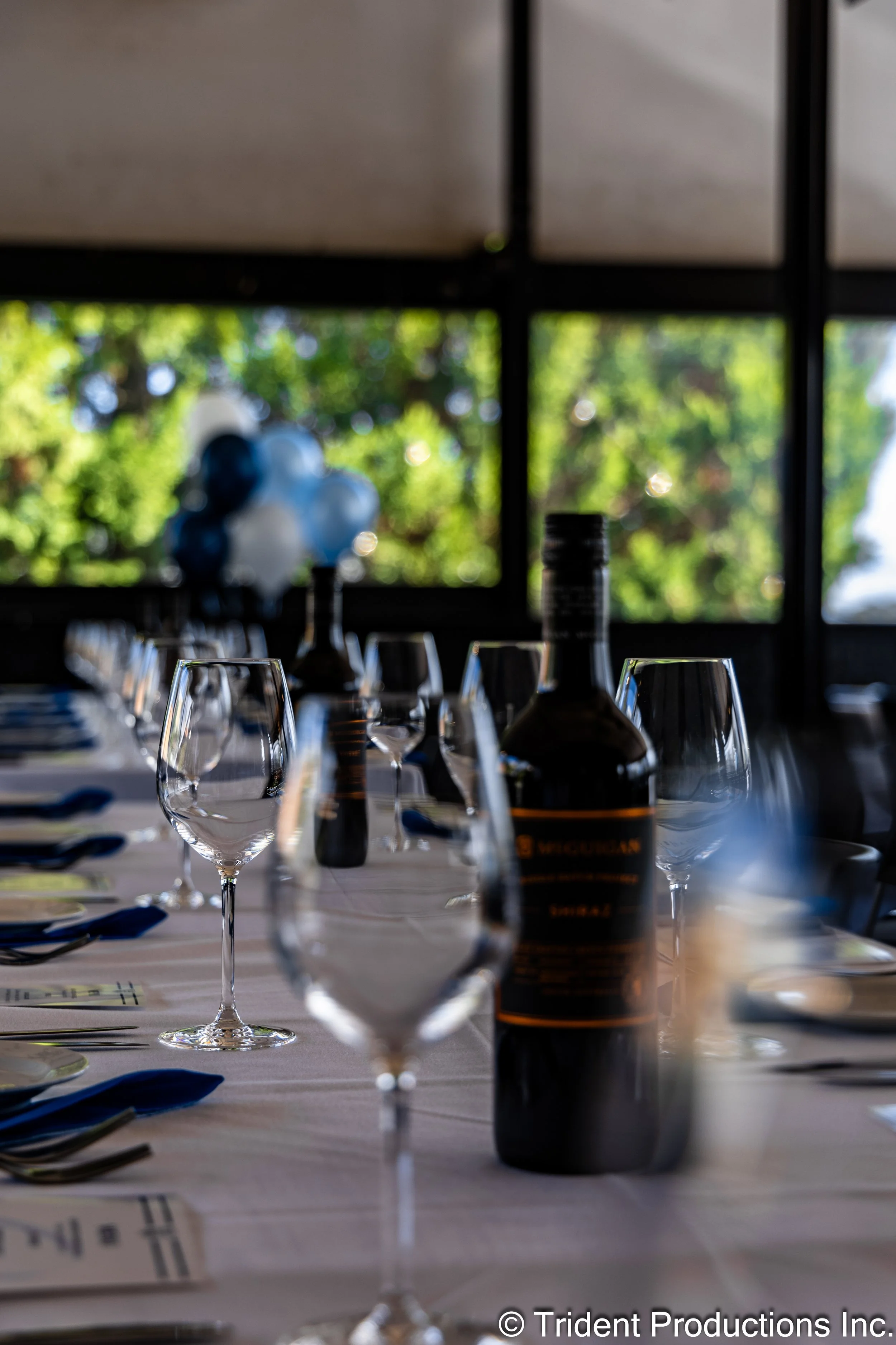 A table set for a formal event with wine glasses, plates, and utensils; a bottle of wine or champagne and balloons in the background, with large windows and greenery outside.