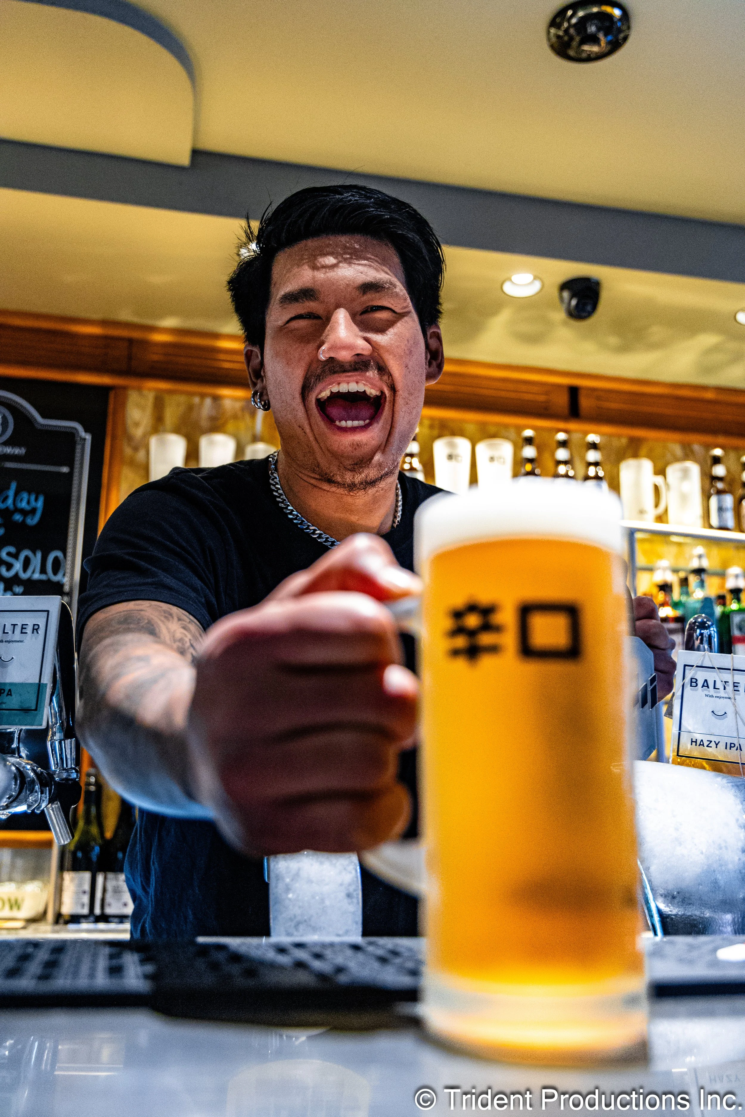 A man with black hair, tattoos, and earrings is smiling and holding a large mug of beer towards the camera in a bar.