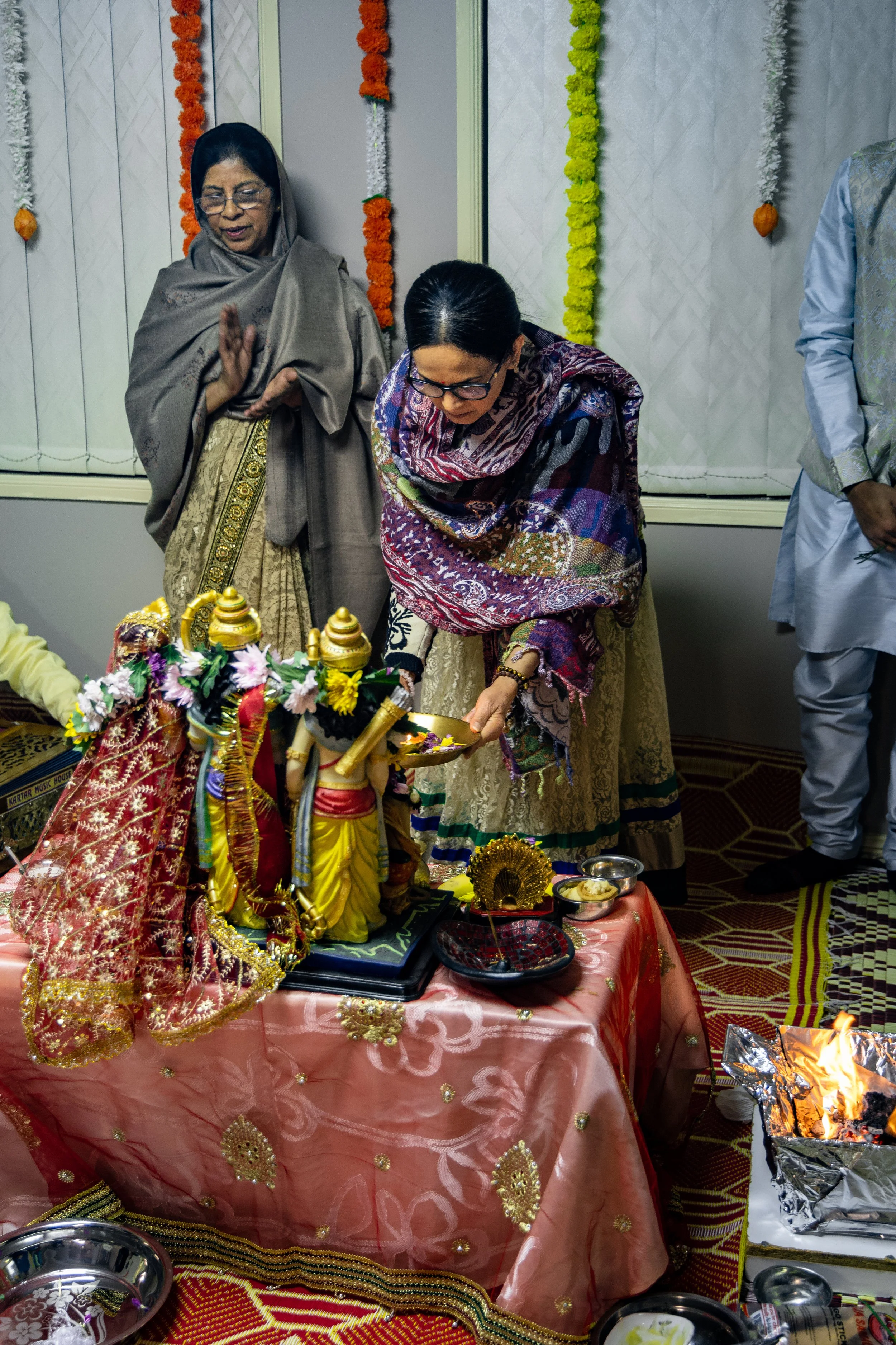 Two women dressed in traditional Indian attire performing a religious ritual with statues of Hindu deities, surrounding items, and a small fire on the side.