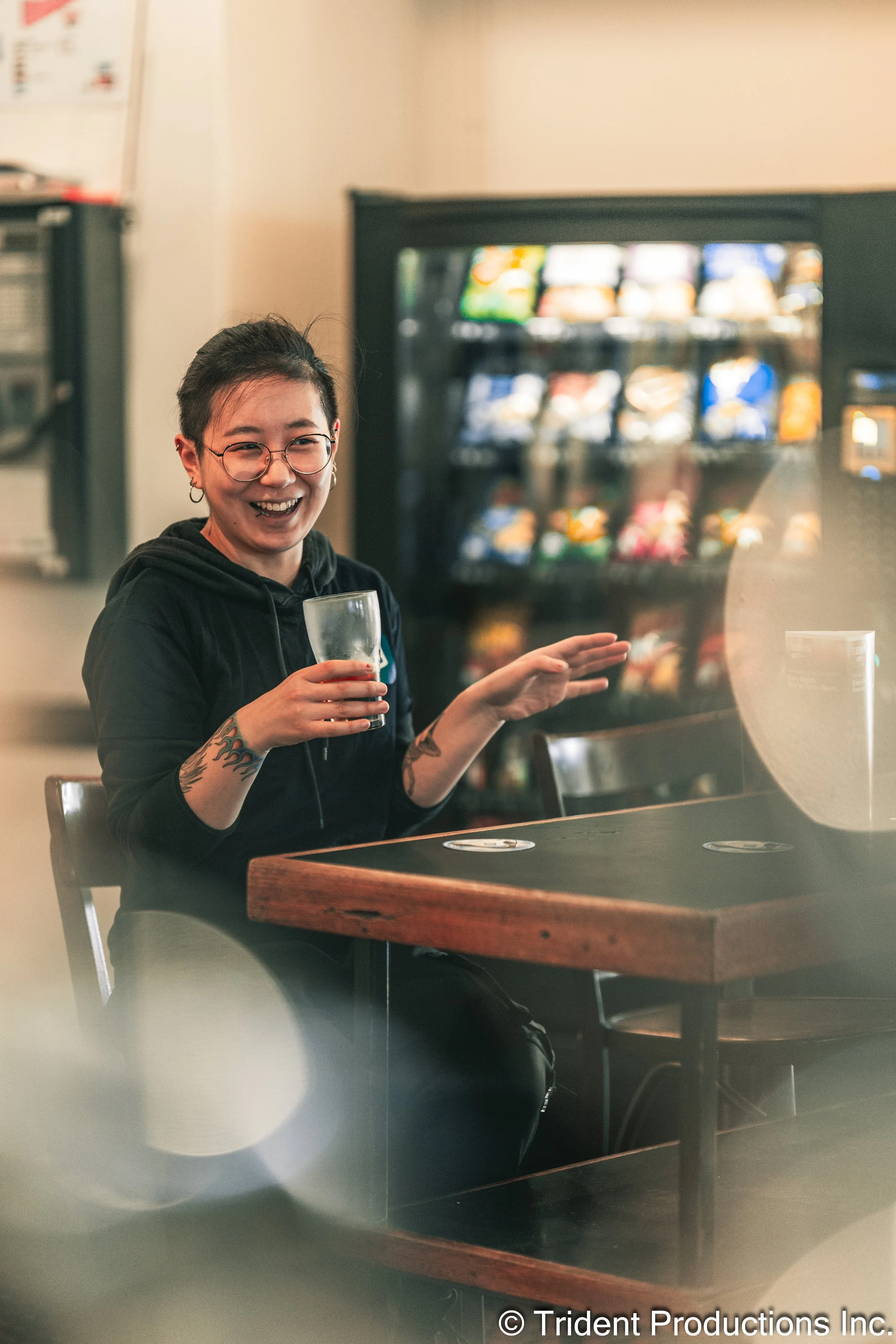 A woman with short dark hair, glasses, and tattoos on her arms, wearing a black hoodie, sitting at a wooden table in a casual setting, holding a clear glass, smiling while engaging in conversation.