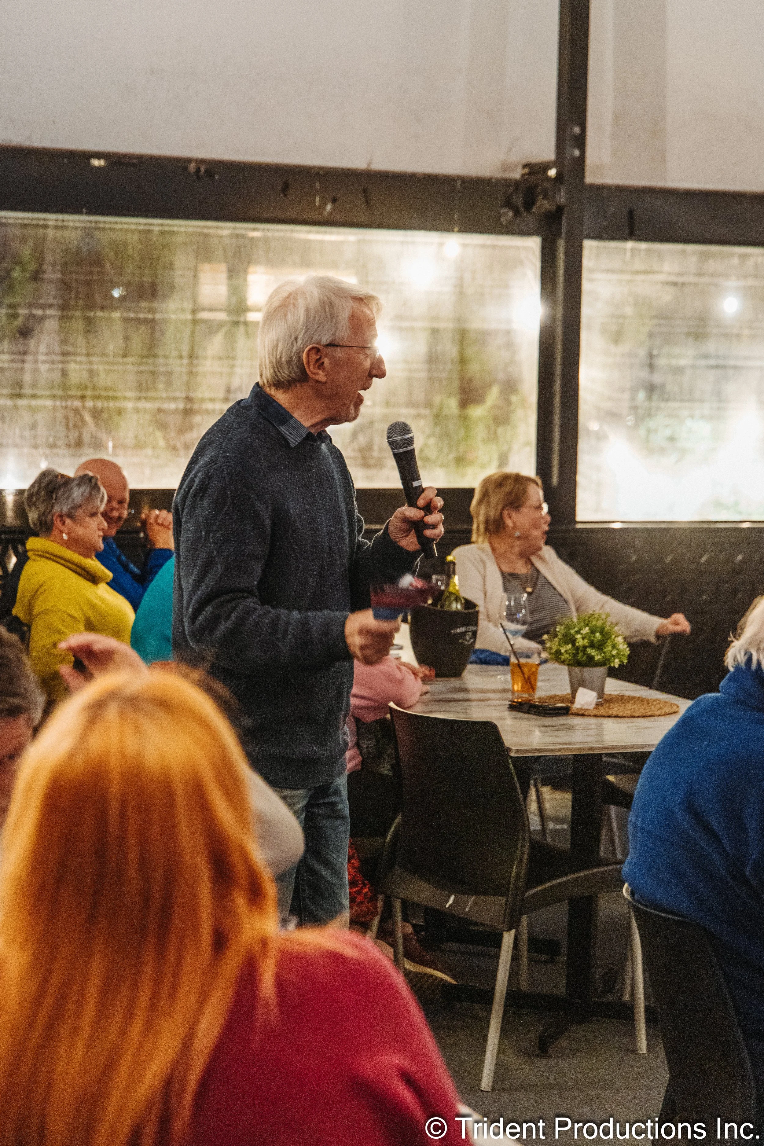 An older man holding a microphone and a small object in his hand, speaking at a gathering in an indoor setting with several seated people listening.