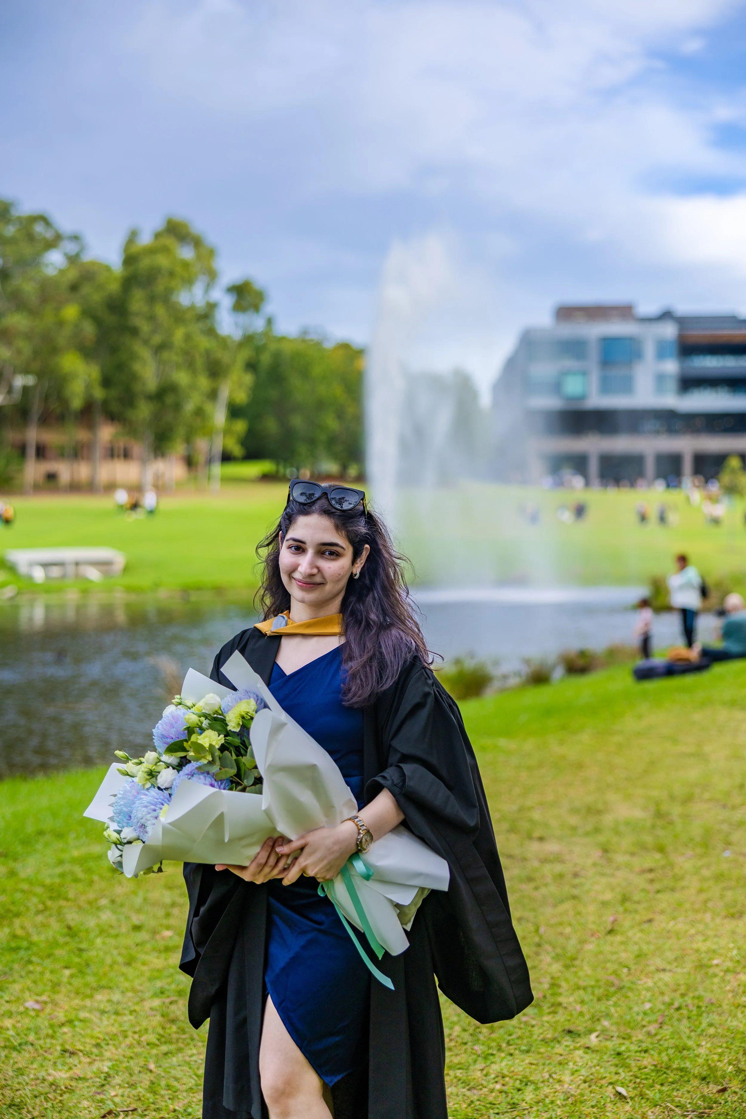 Young woman in a graduation gown and cap holding a bouquet of flowers in a park with a fountain, trees, and modern buildings in the background.