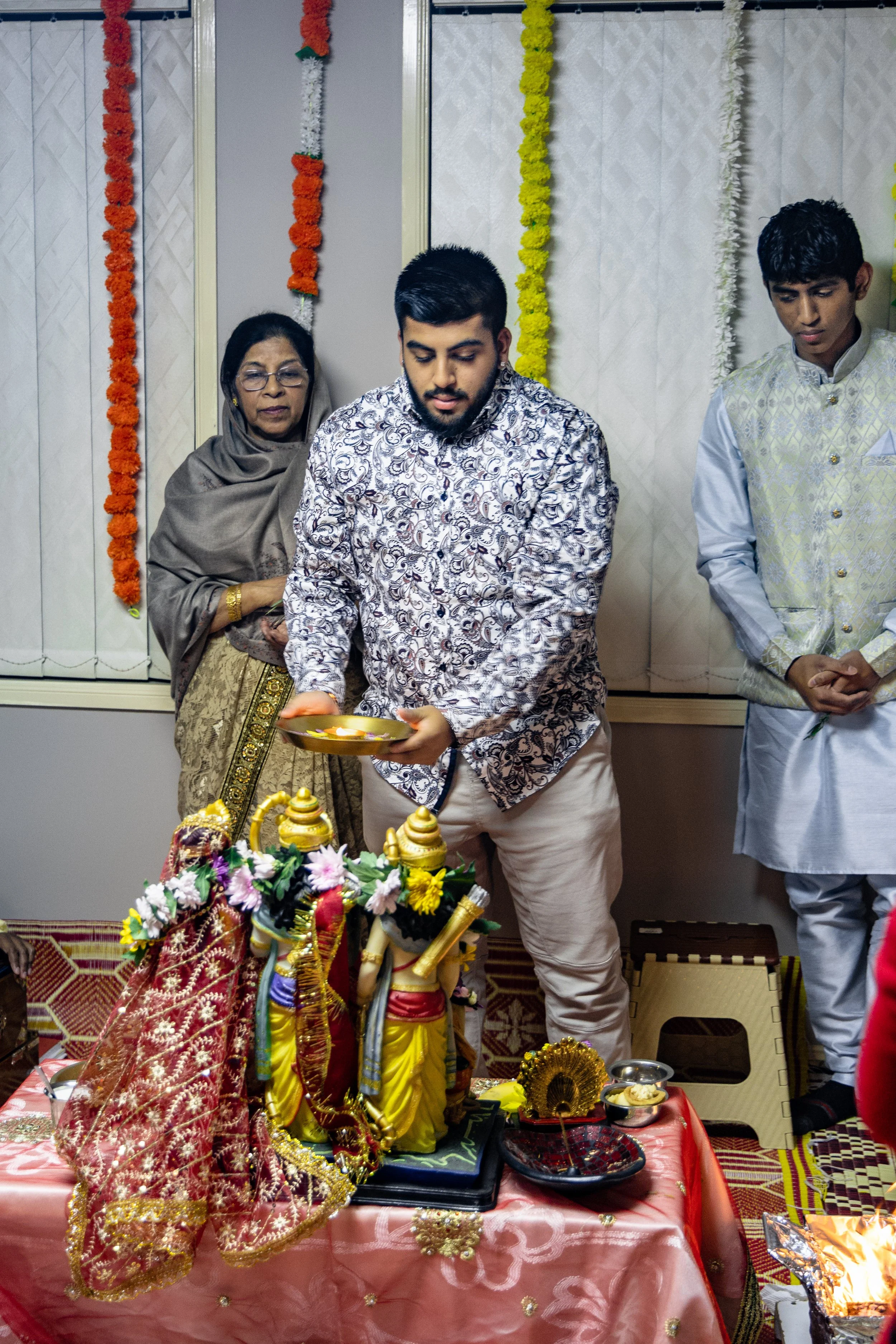 People participating in a Hindu religious ceremony with idols, flowers, and offerings on a decorated table.
