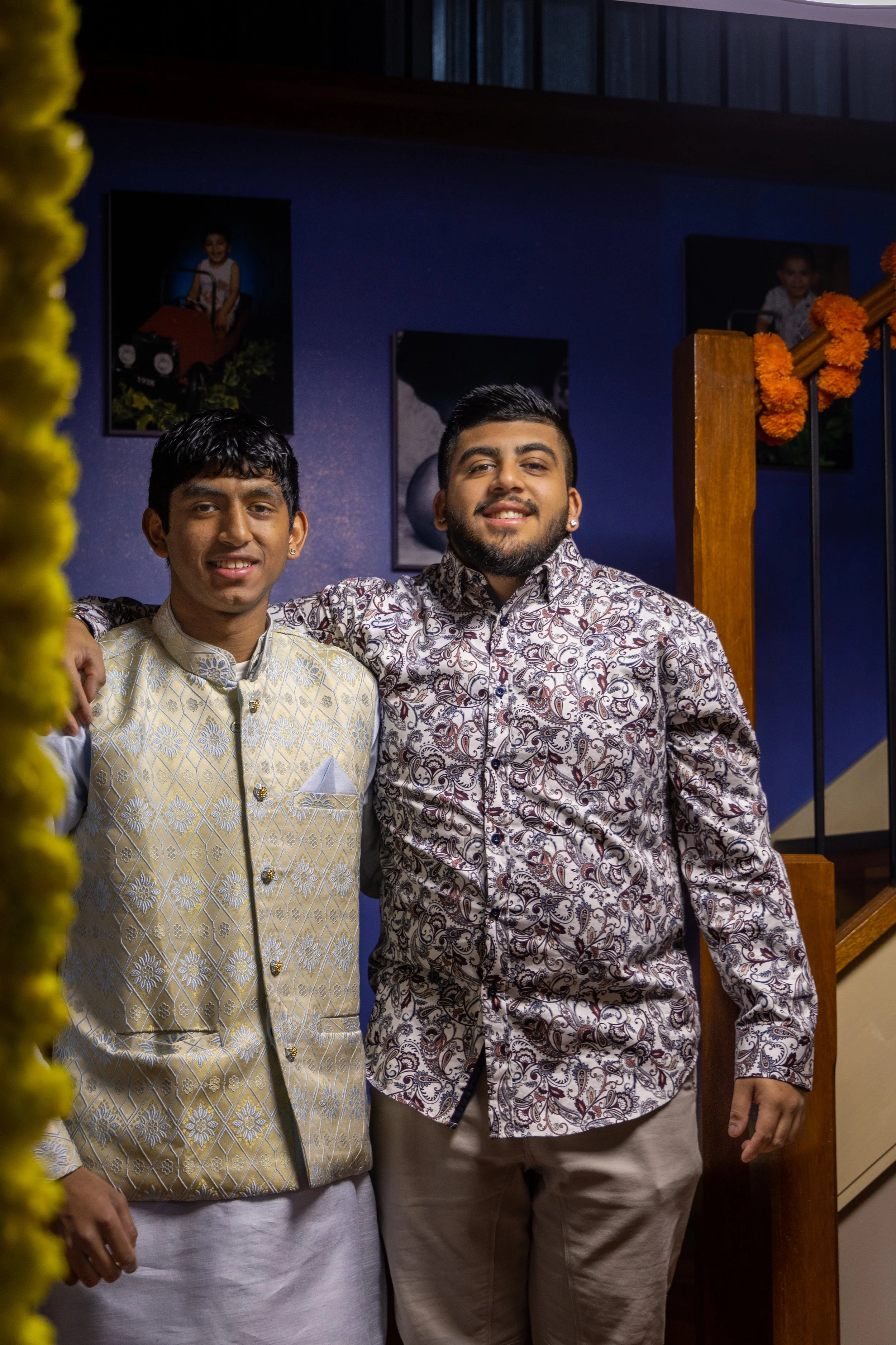 Two men standing indoors near a staircase, smiling at the camera, with framed photographs on a blue wall in the background.