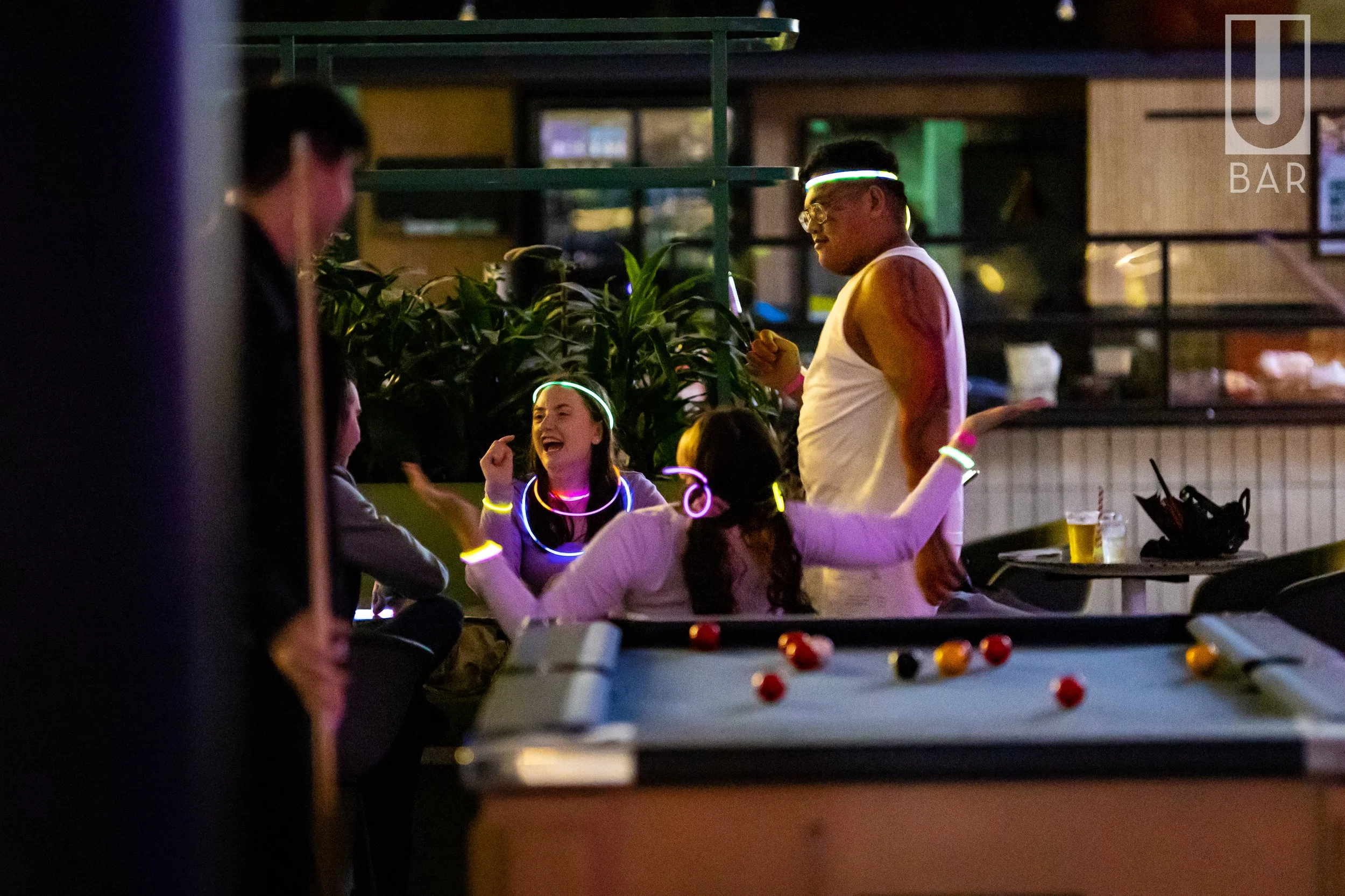 People enjoying a night at a bar with glow-in-the-dark accessories, playing pool, and having drinks.