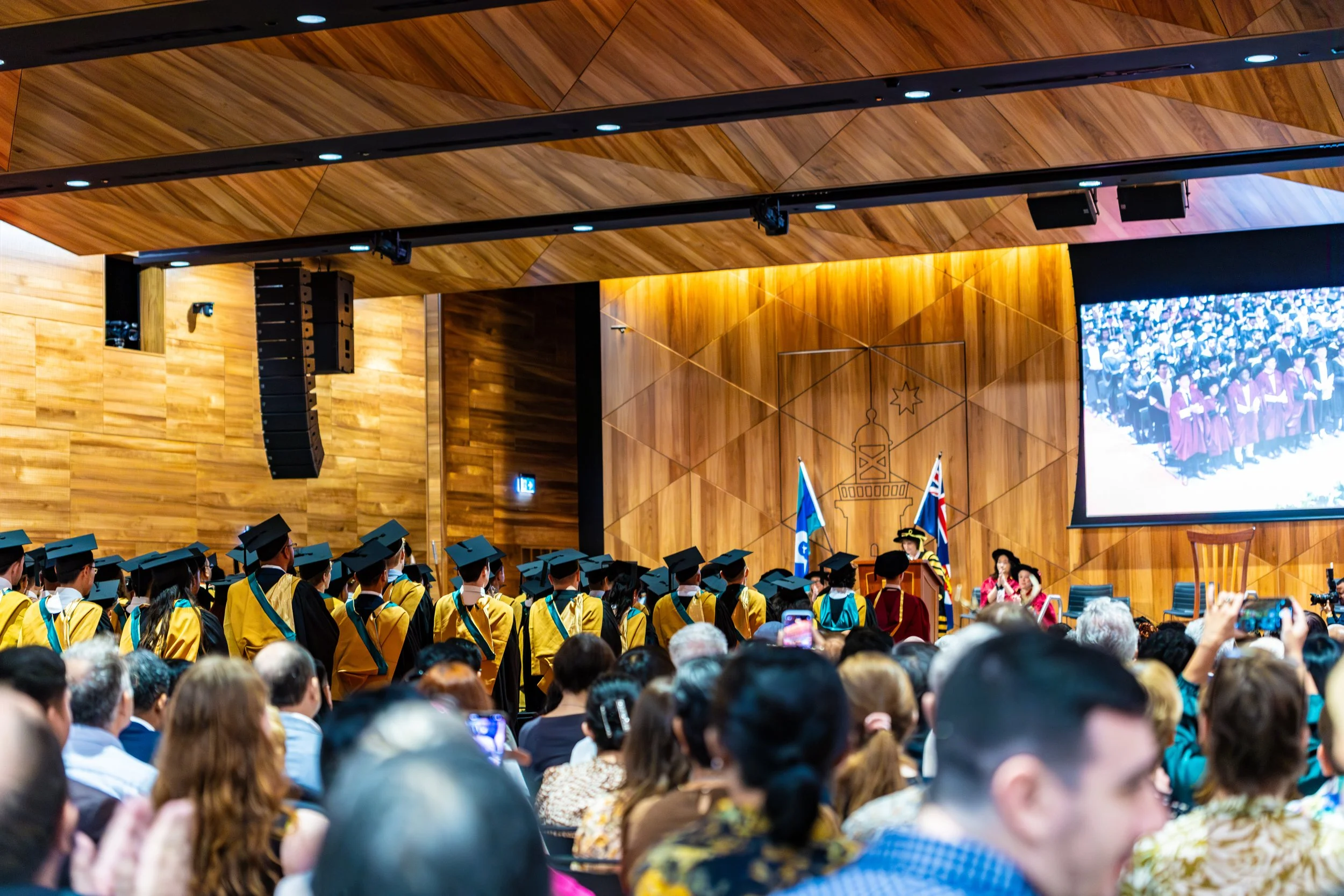 An indoor graduation ceremony with graduates in caps and gowns standing on stage in front of an audience, with flags and a large screen displaying a photo of graduates.