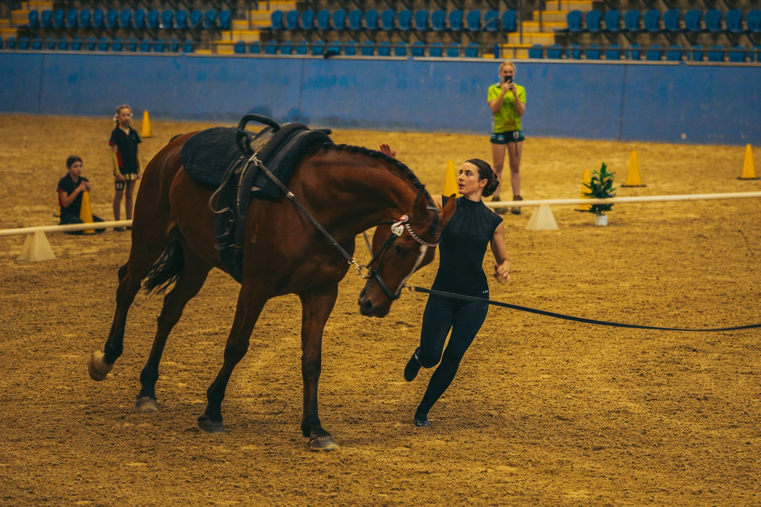 A womanleading a brown horse in an indoor equestrian arena, with spectators and children watching in the background.
