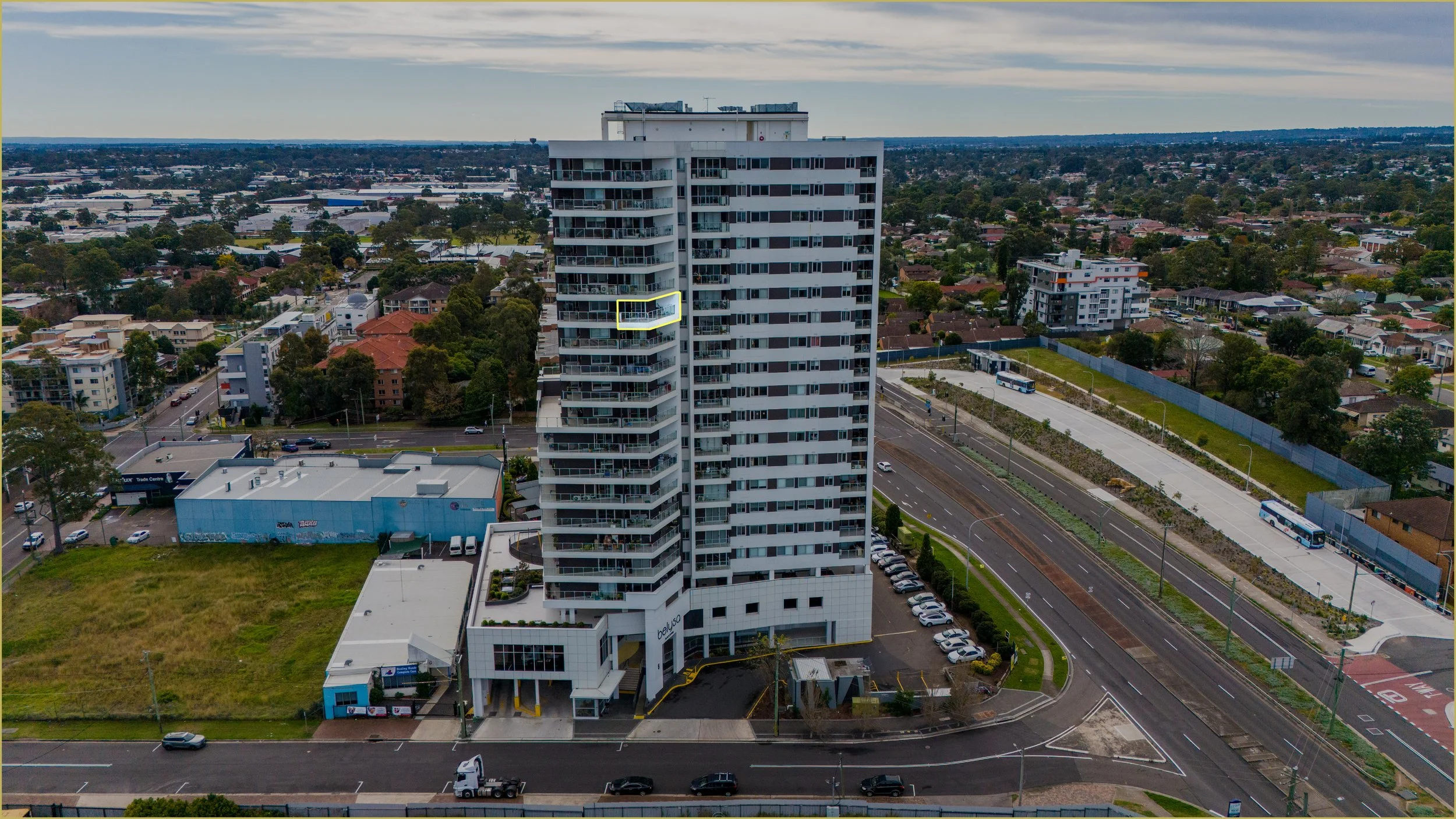 An aerial view of a tall white apartment building with balconies, surrounded by a parking lot, adjacent to a bus stop, with residential houses and trees in the background.