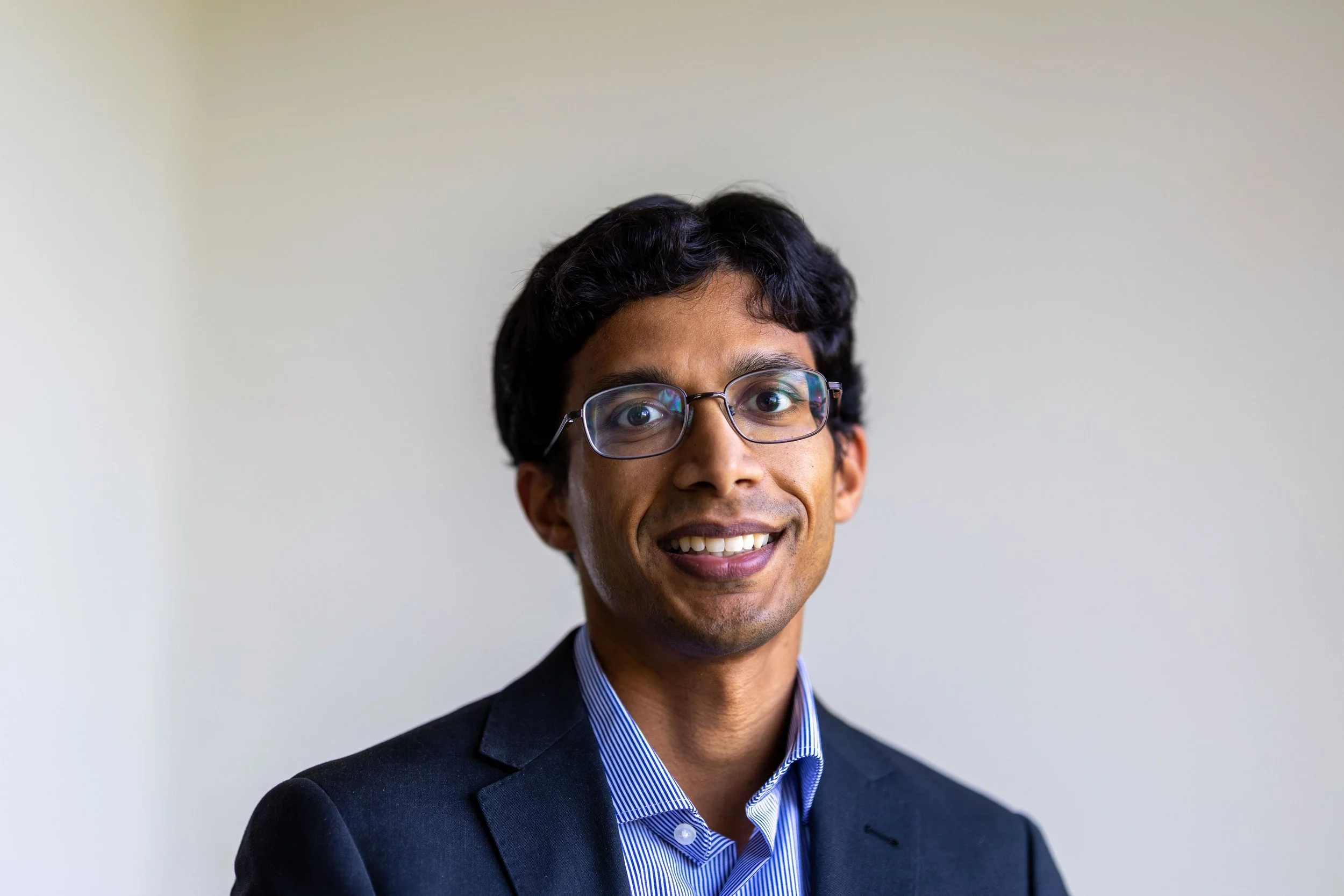 Portrait of a smiling man with glasses wearing a dark blazer and blue striped shirt against a plain background.