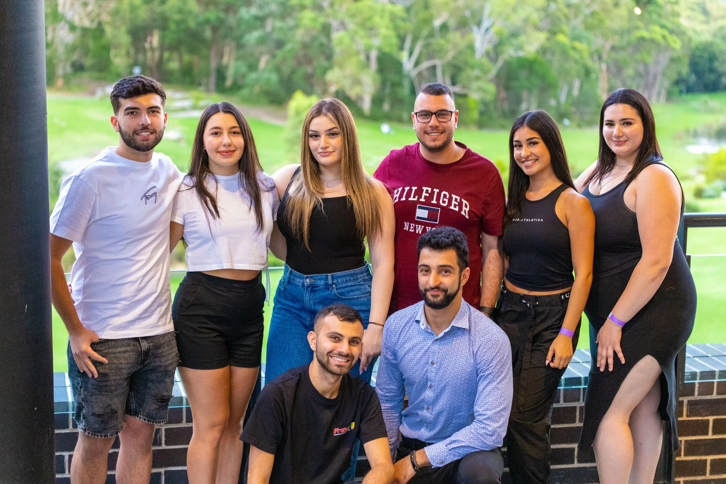 Group of nine diverse young adults posing outdoors on a balcony with a lush green park in the background.