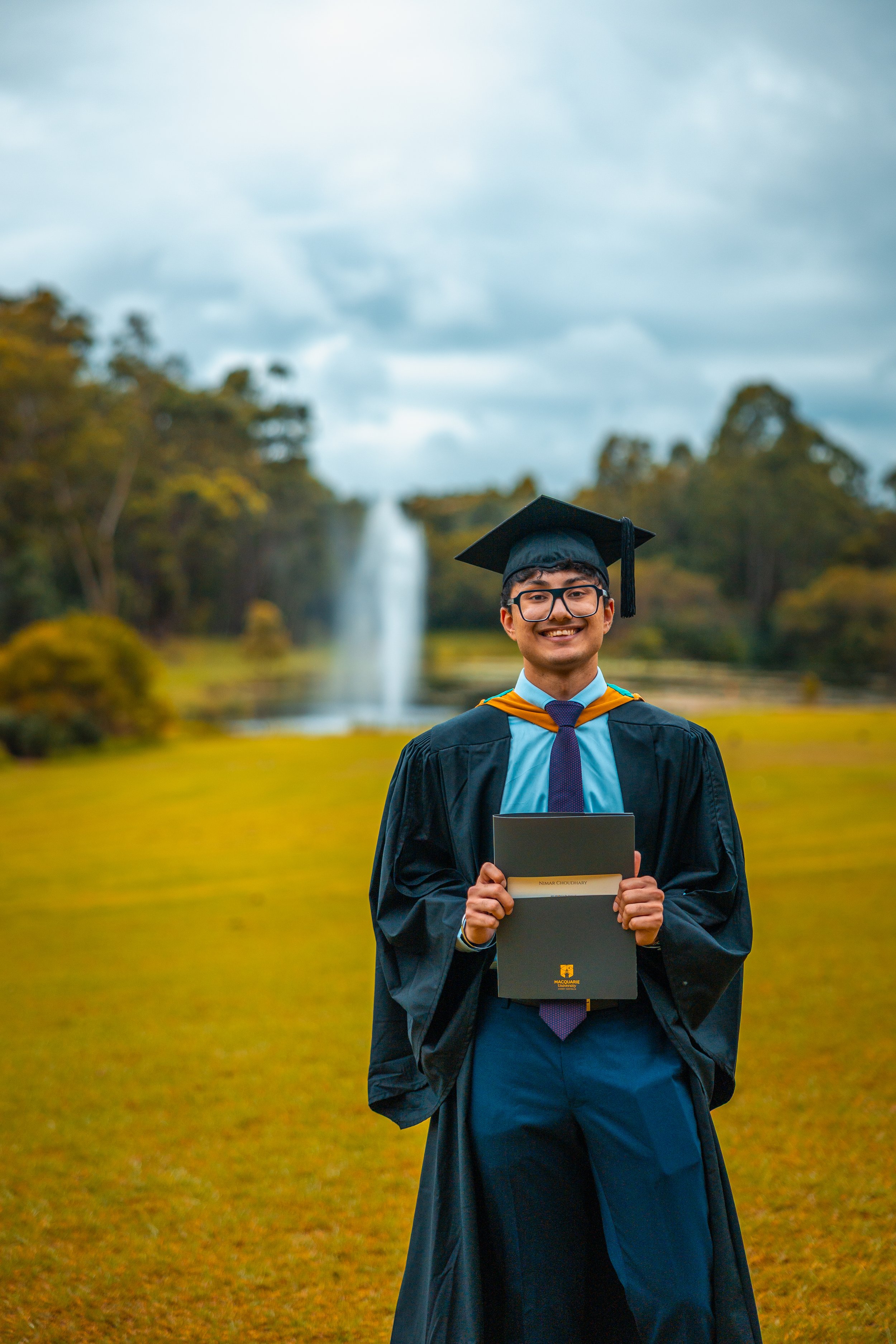 A young man in graduation gown and cap holding a diploma stands outdoors on a grassy field with trees and a waterfall in the background.