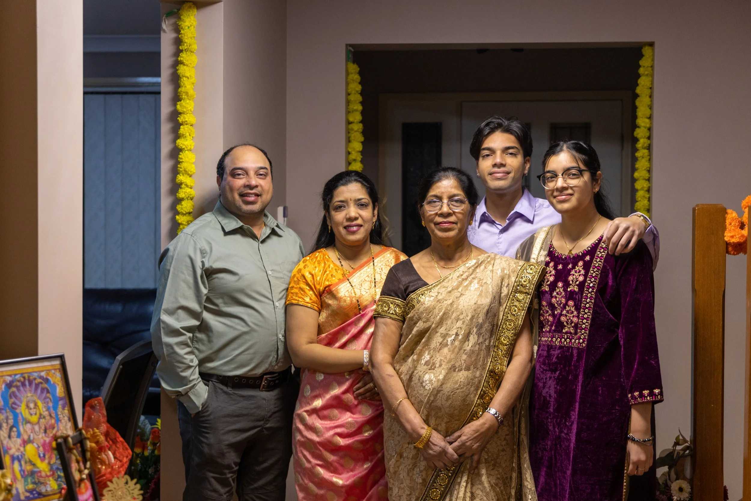 A multi-generational Indian family standing together indoors, dressed in traditional Indian attire, celebrating a festive occasion with yellow marigold garlands decorating the background.
