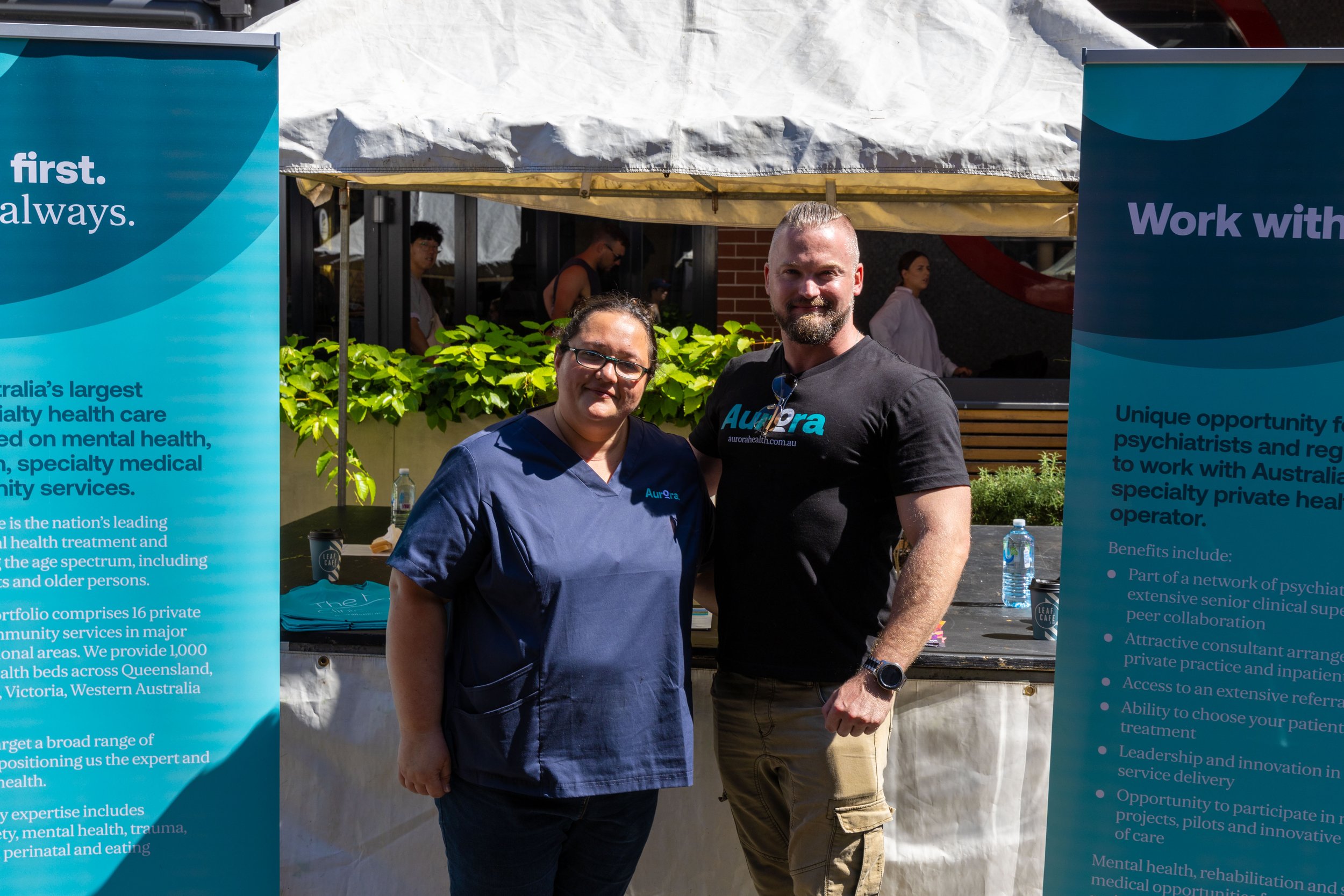 Two people standing outdoors at a booth with blue banners, one wearing medical scrubs and the other in a black t-shirt with 'Aurora' logo, surrounded by trees and other people.