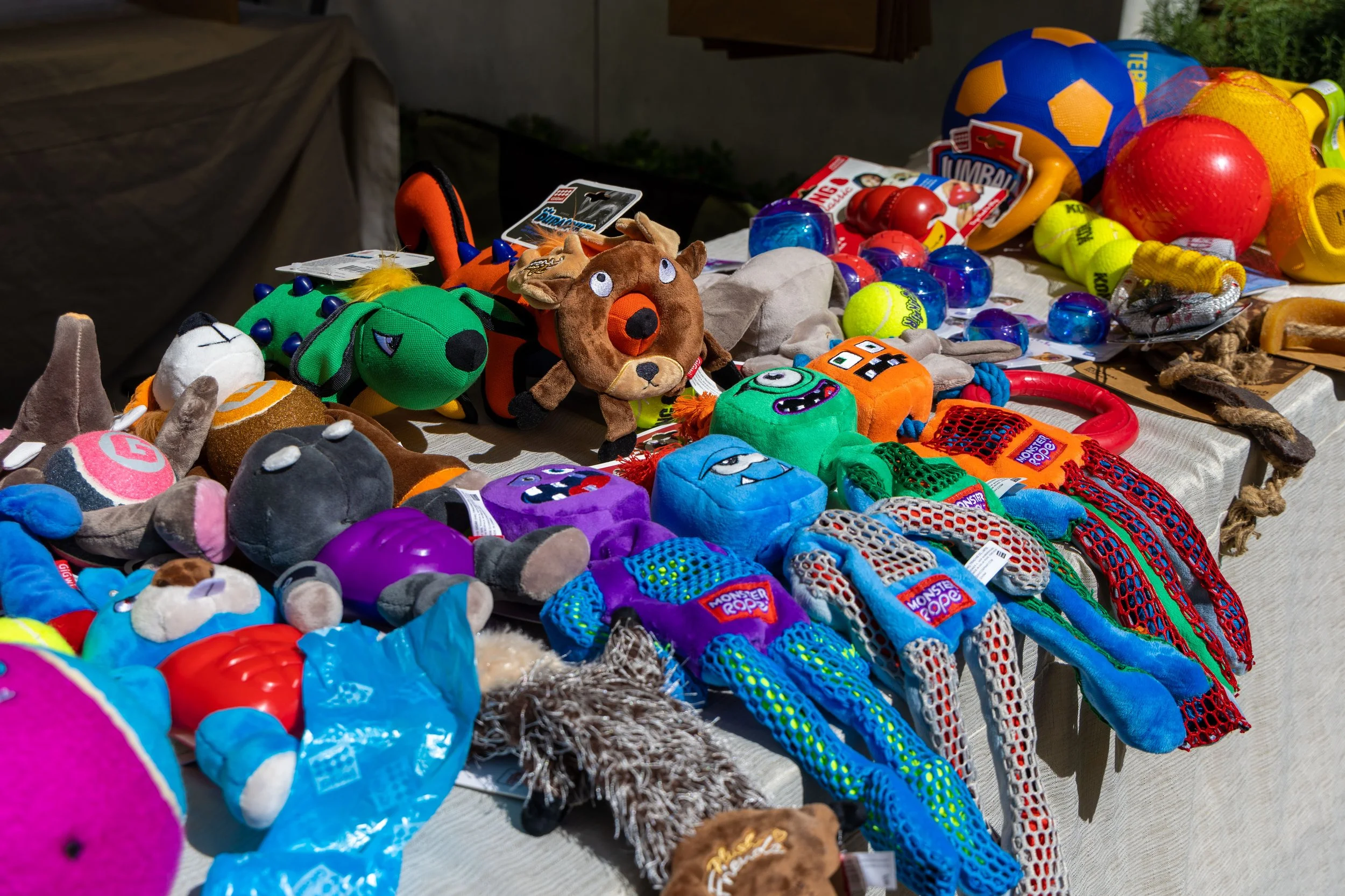 Table with various plush toys, balls, and sports equipment such as tennis balls, soccer balls, and a volleyball, arranged outdoors.
