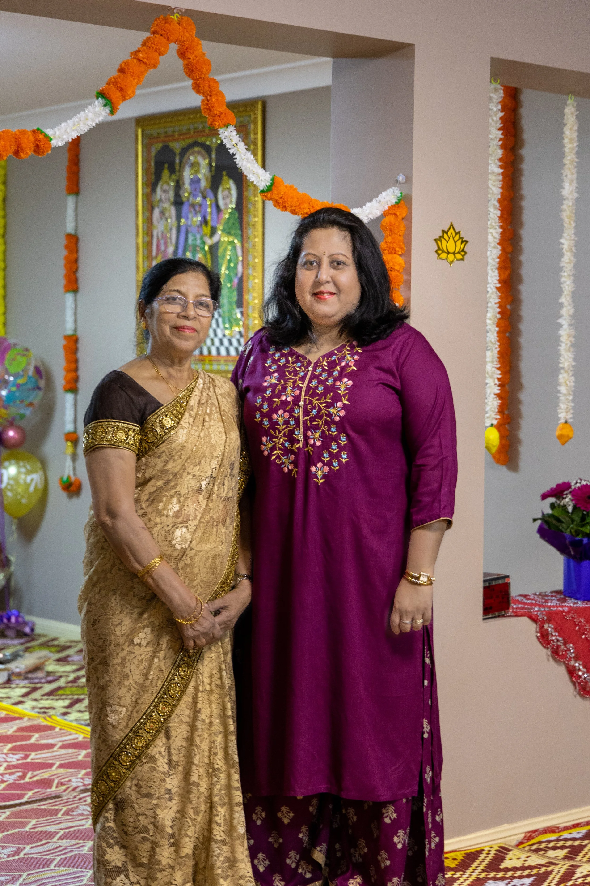 Two women dressed in traditional Indian attire standing together at a celebration or festival, decorated with marigold flower garlands and balloons.