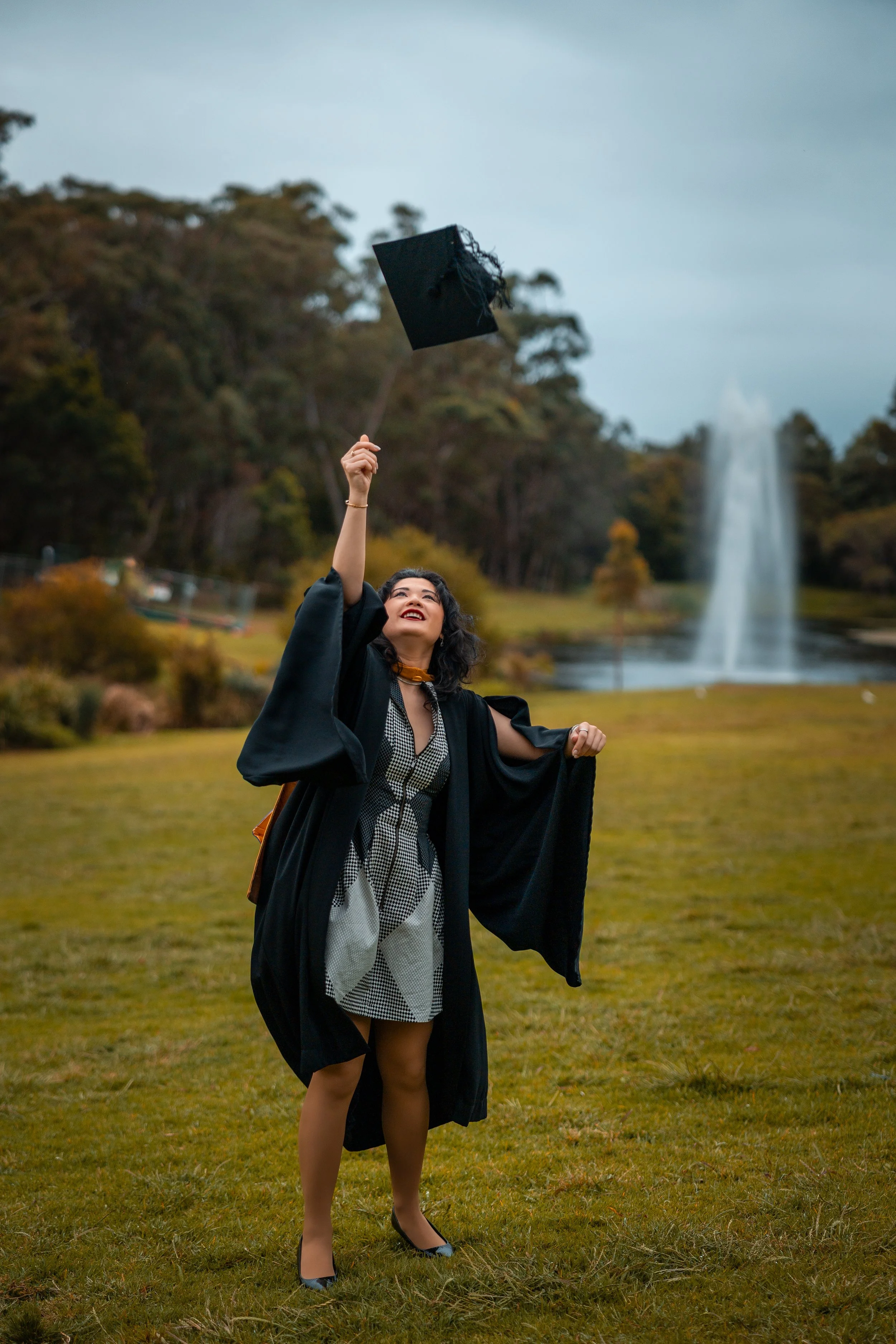 A woman in a graduation gown and cap throws her cap into the air outdoors in a park with a fountain in the background.