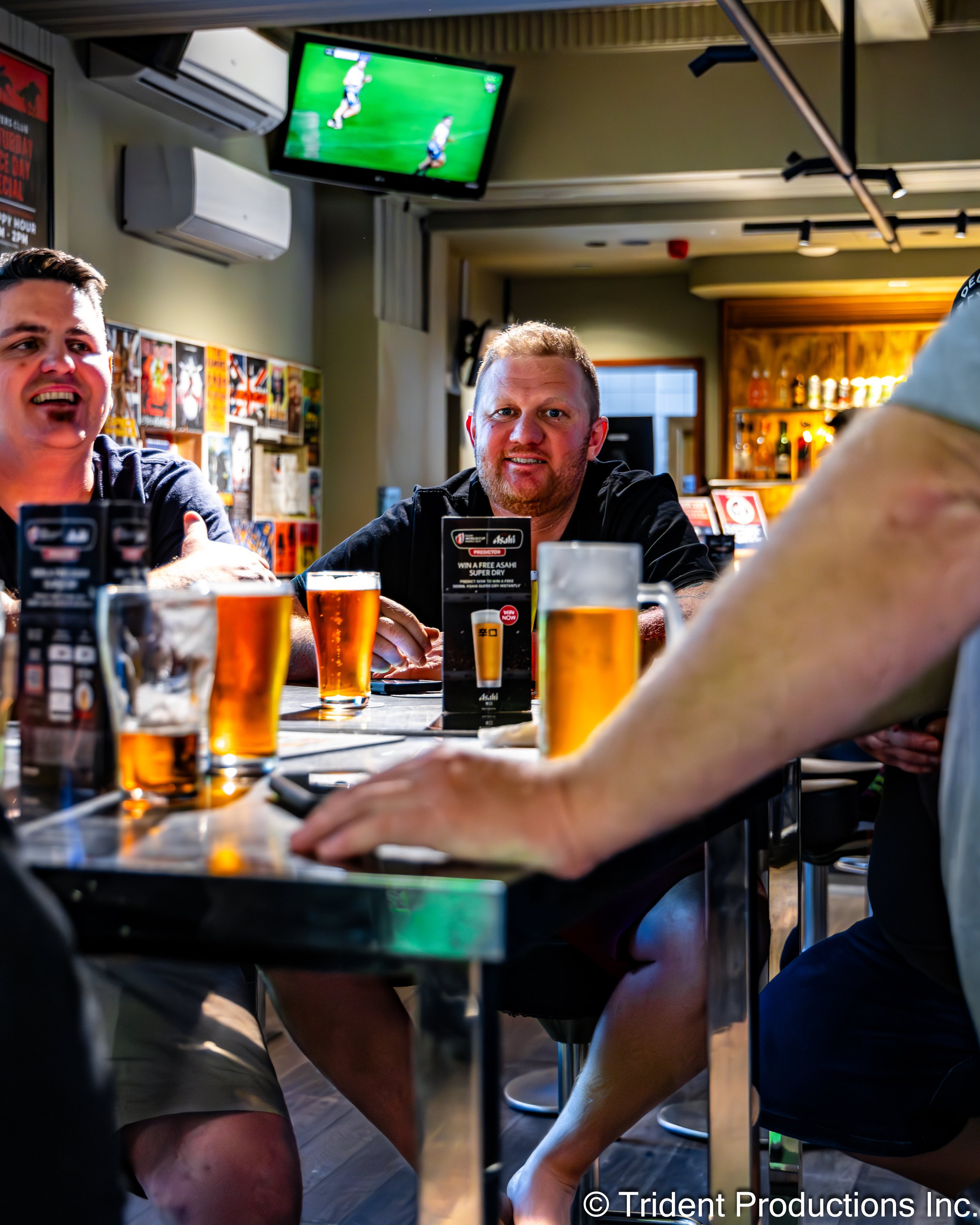 People sitting at a bar table with glasses of beer, engaged in conversation, with a TV showing a soccer game in the background.