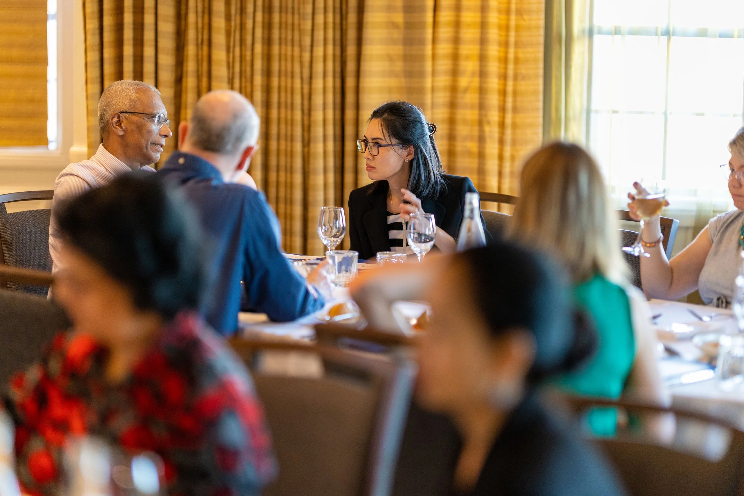 Group of diverse people having a discussion at a formal dining table in a room with yellow curtains.