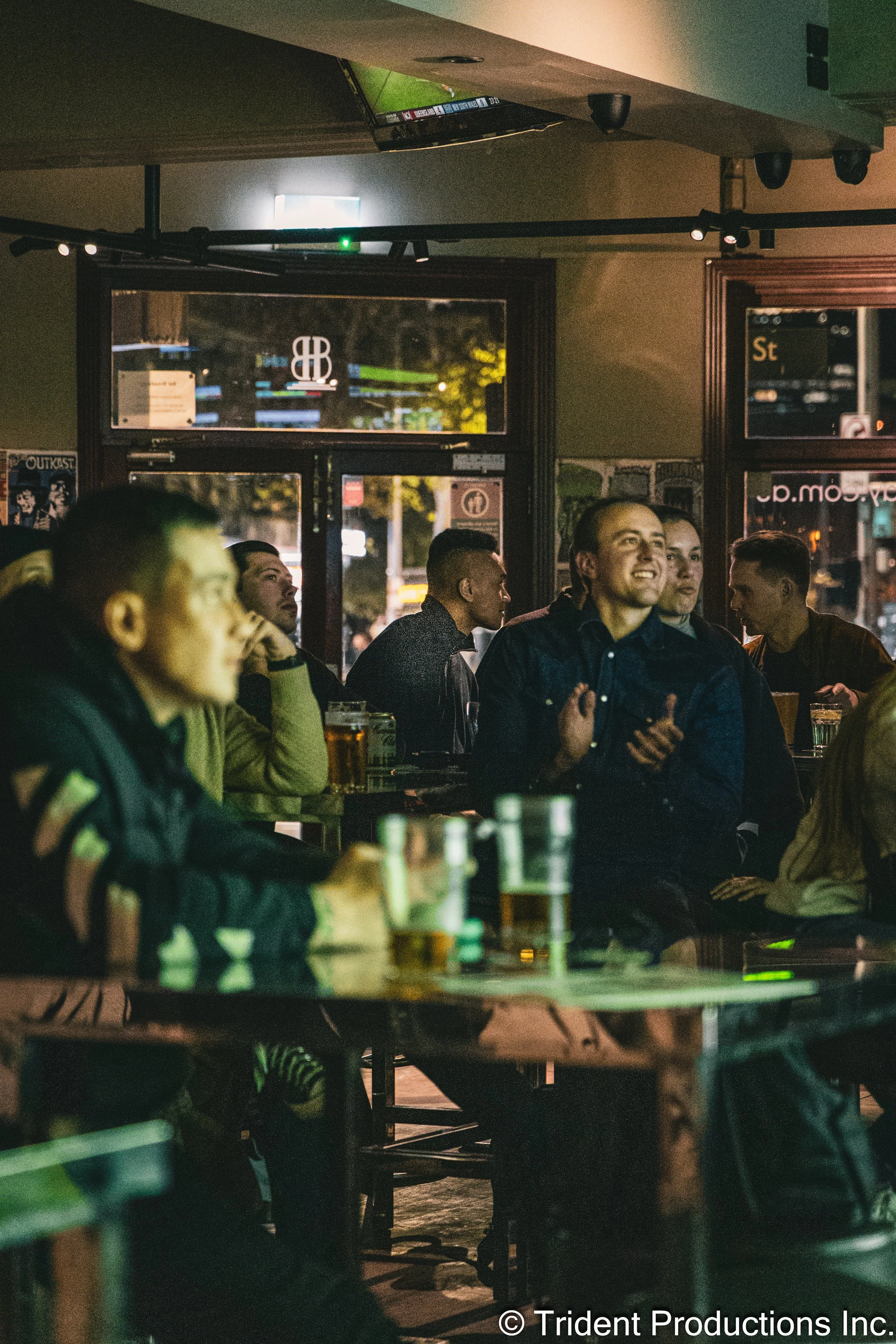 People sitting inside a bar or restaurant, some watching a sports game on a TV, with drinks on the tables, in an urban setting at night.