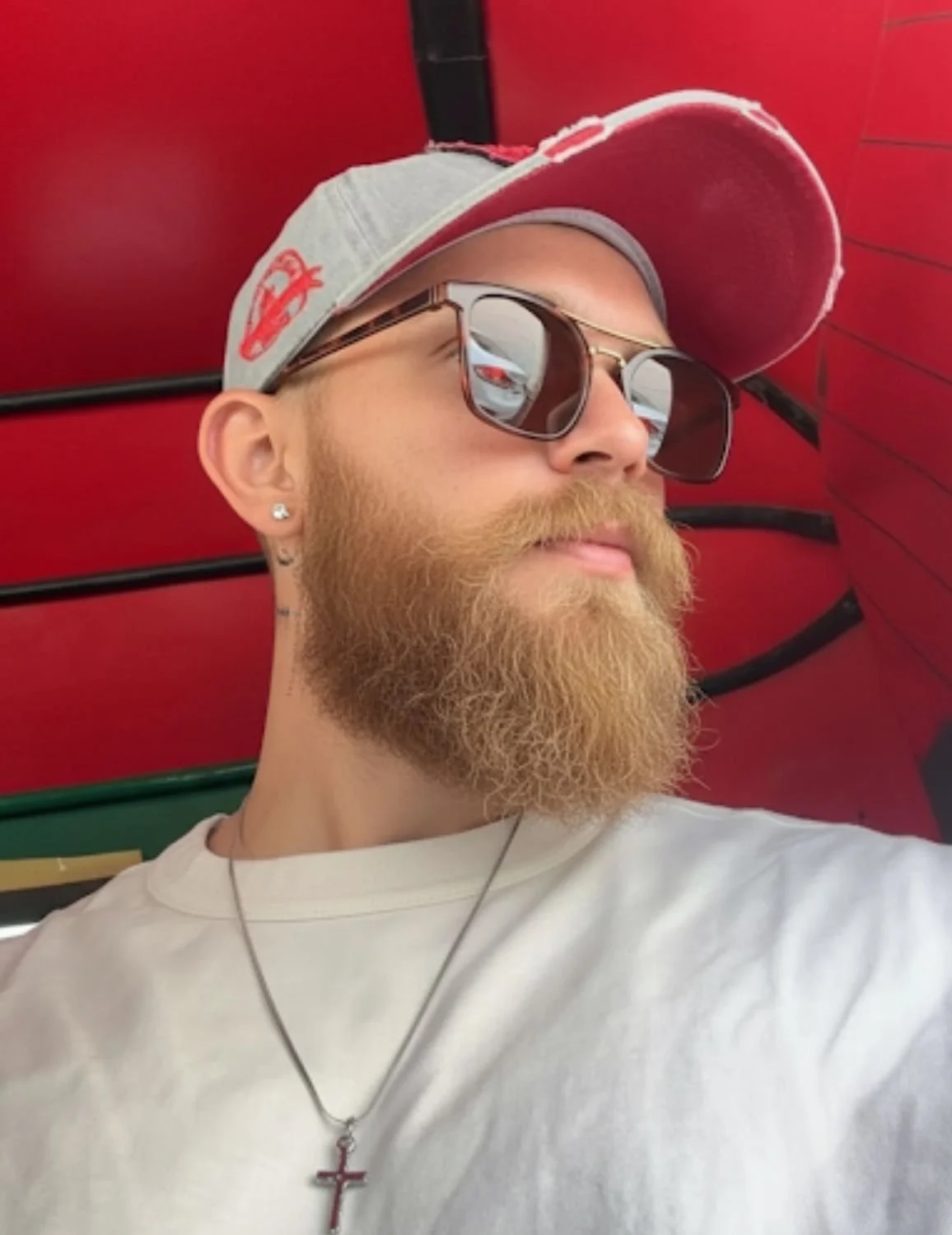A man with a red beard and mustache wearing sunglasses, a baseball cap, a white T-shirt, and a cross necklace, with a red umbrella in the background.