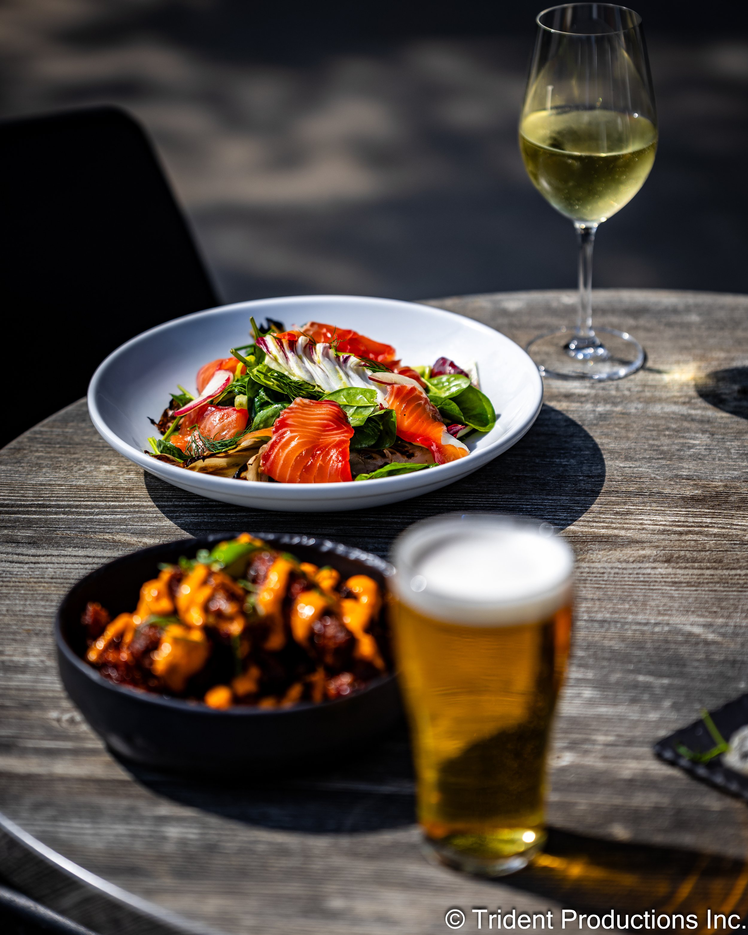 A wooden table with a glass of white wine, a bowl of salad with smoked salmon and greens, a bowl of glazed meatball dish, and a glass of beer.