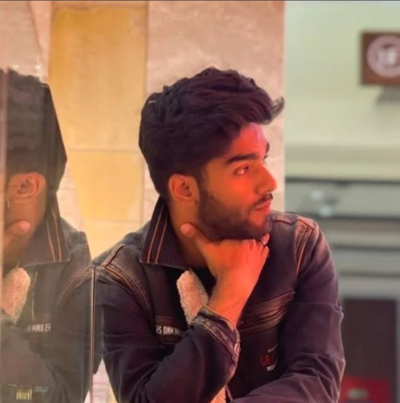 Young man with dark hair and beard, wearing a dark denim jacket, sitting indoors, resting his chin on his hand, looking to the right. Reflection of him can be seen in a mirror beside him.