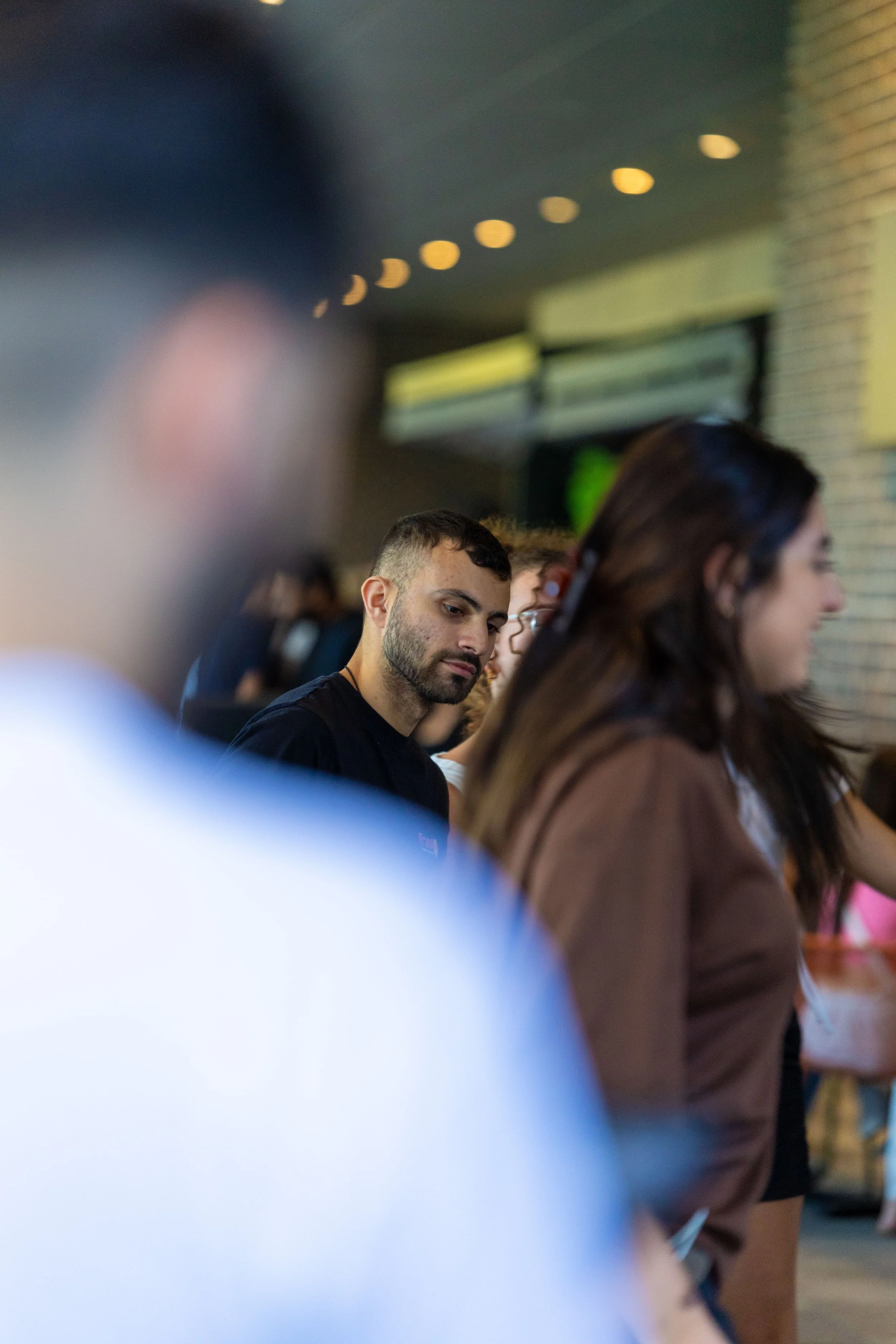 People standing in line inside a building, some looking at their phones, with a man with a beard and black shirt in focus.