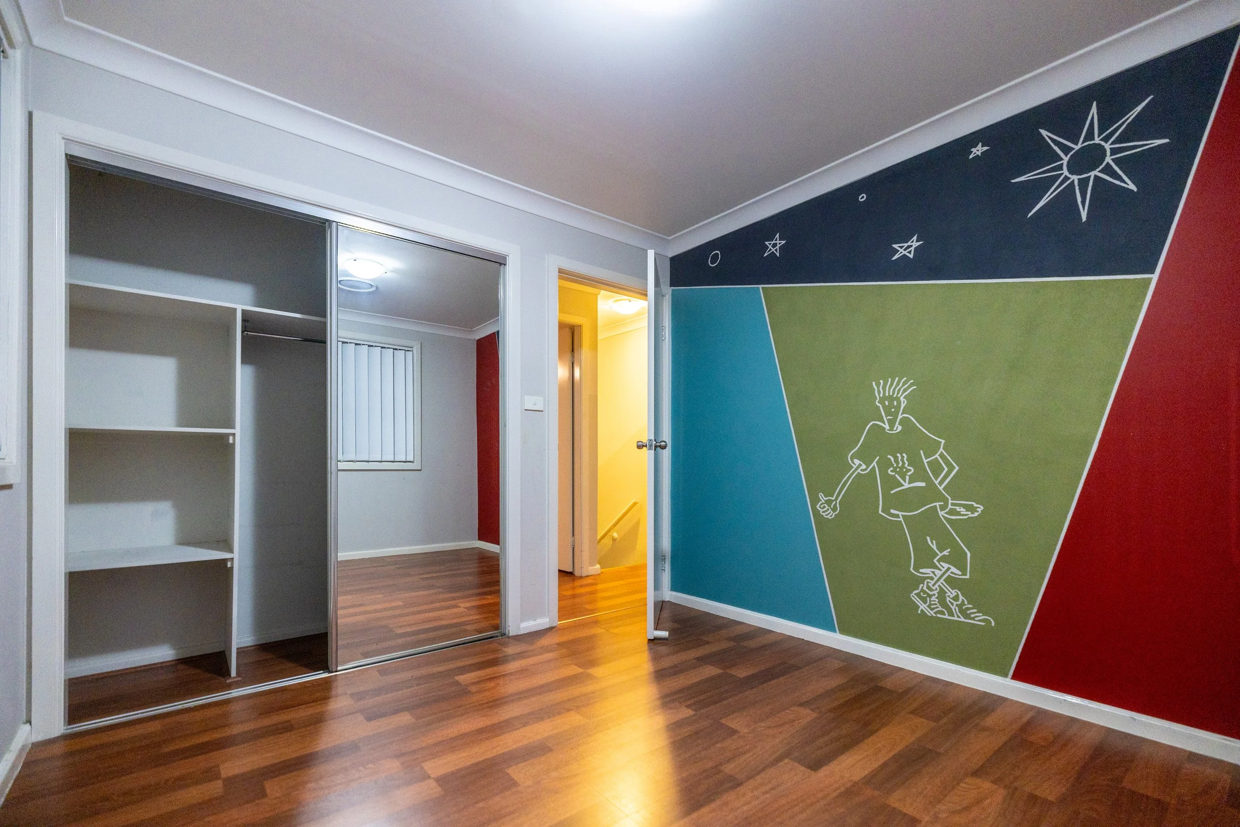 Room with wooden floors, a closet with sliding mirror doors, a colorful wall mural with abstract shapes, stars, and a stylized drawing of a person with spiky hair, wearing casual clothes.