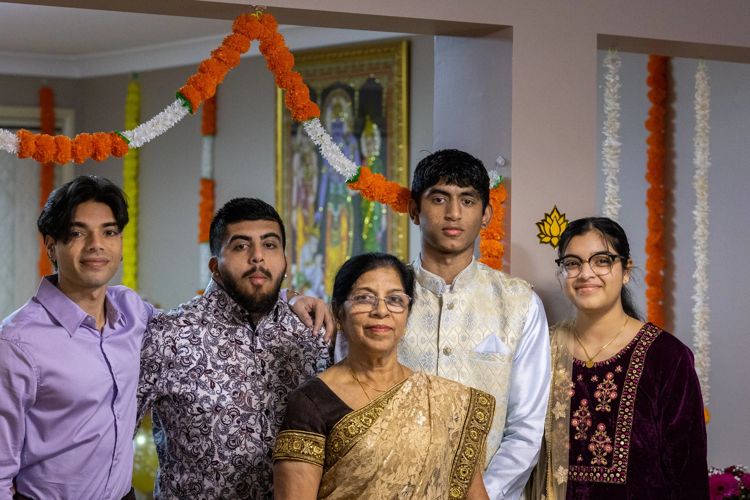 A family of six people dressed in traditional Indian attire standing together indoors, decorated with orange and white garlands for a celebration.