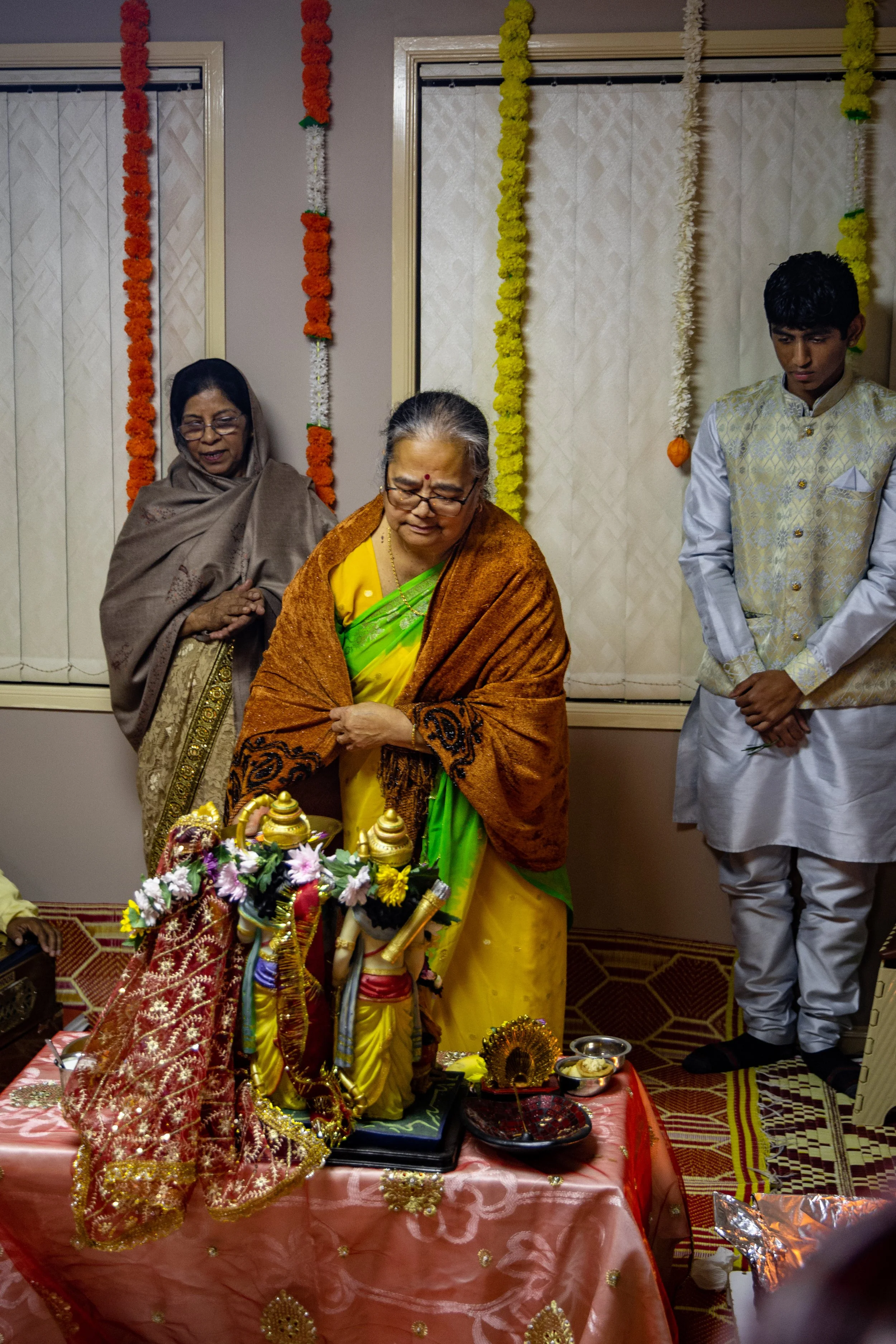 Three people, two women and one young man, participate in a traditional Indian religious ritual. The women are wearing sarees, with one in yellow and orange and the other in brown, while the young man wears a cream-colored kurta. They stand around a 