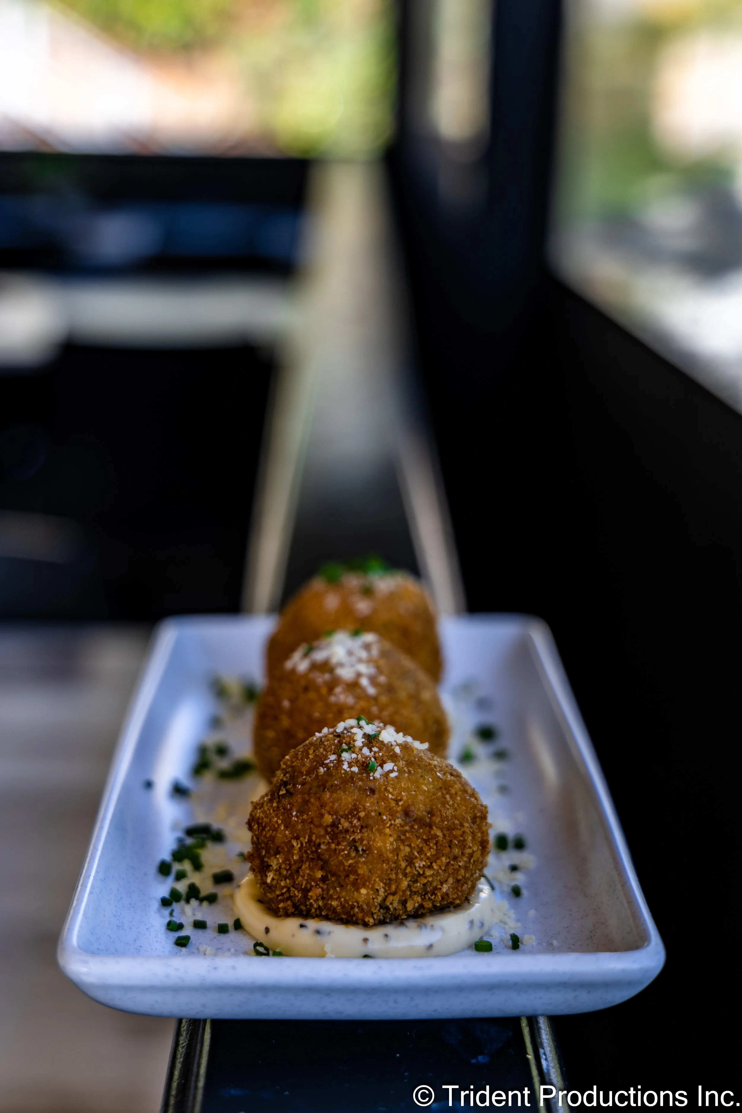 Three breaded and fried arancini served on a white rectangular plate, garnished with chopped chives and a dollop of creamy sauce, with a blurred background of a window and interior.