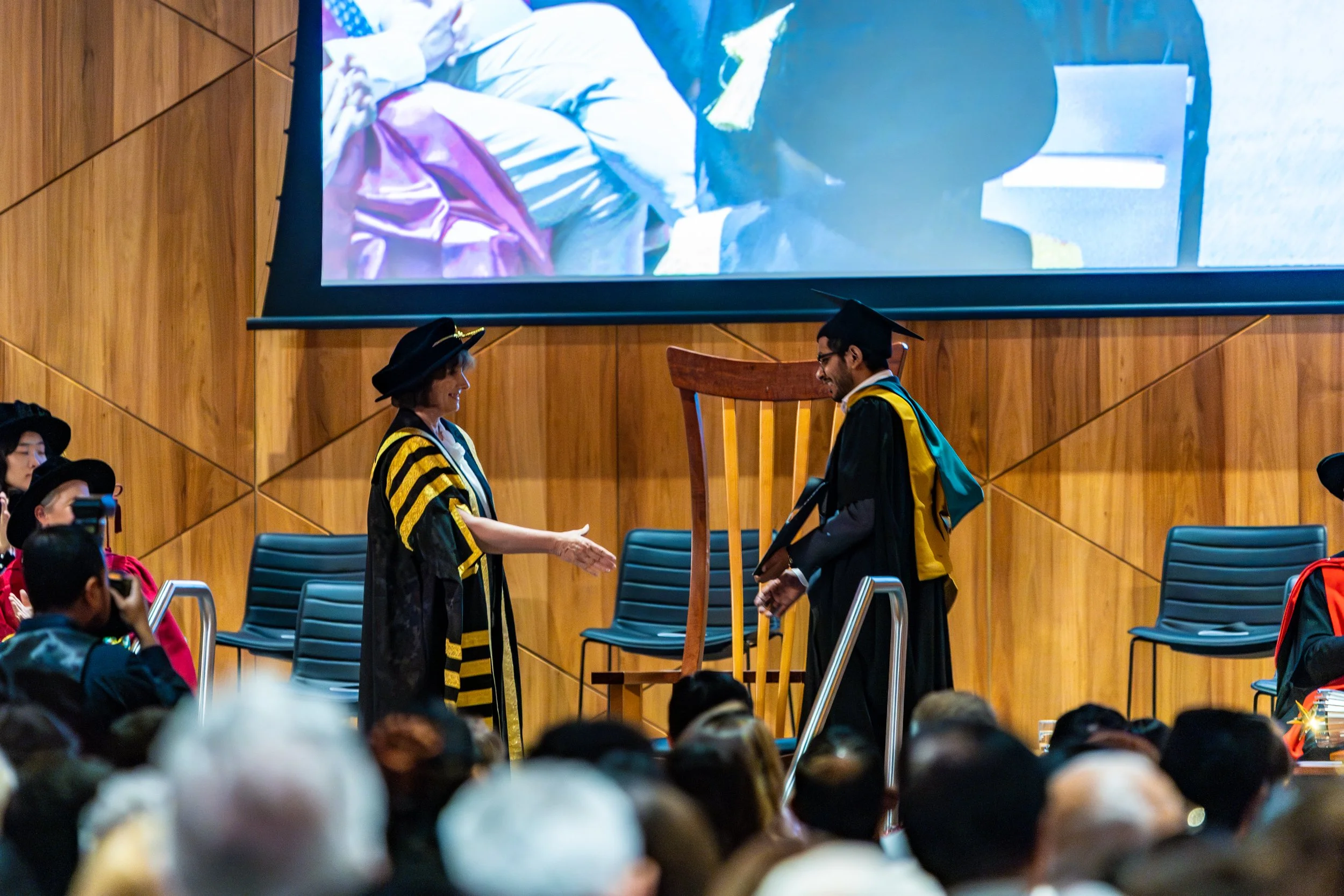 A graduation ceremony with a woman in academic regalia shaking hands with a young man in cap and gown on stage, with an audience in the foreground and a large projection screen behind them.