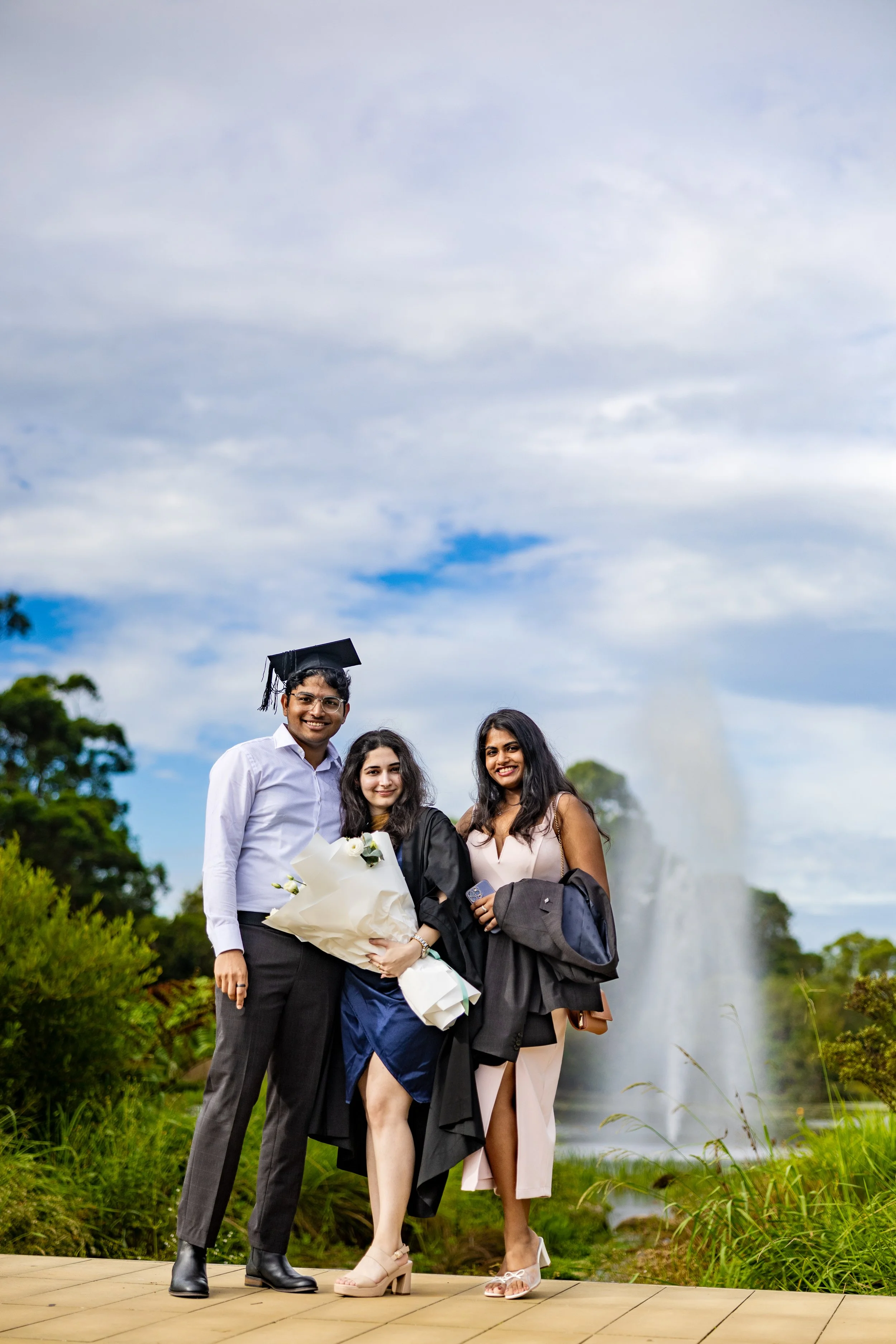 Three young adults celebrating a graduation outdoors, one wearing a cap and gown, holding a bouquet, with a fountain and greenery in the background.
